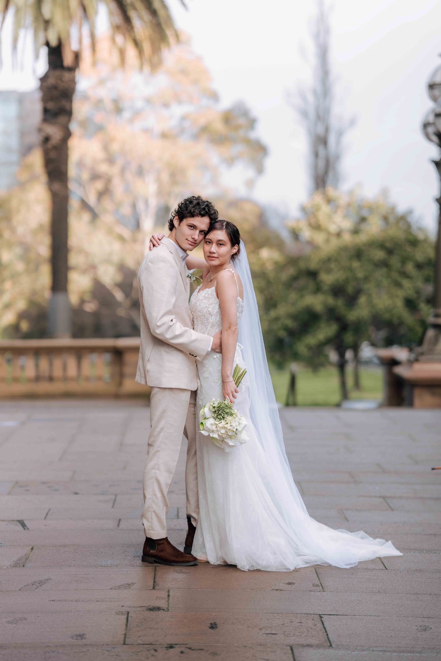 Bride and groom embrace warmly on stone terrace with palm trees, she holds white bouquet in lace gown with veil.