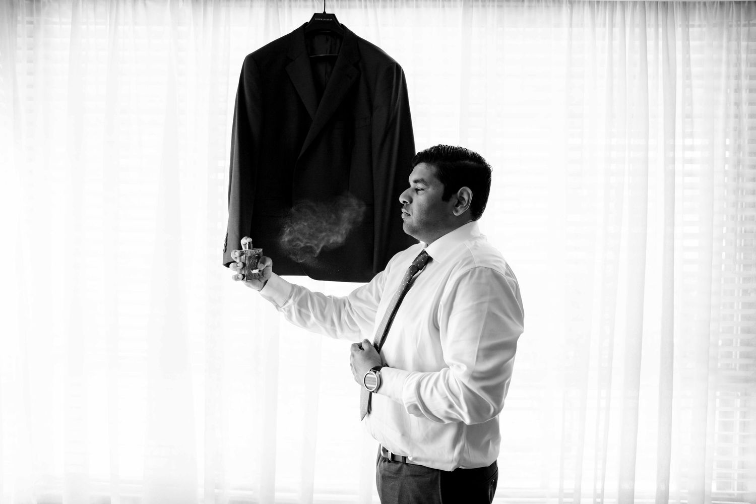 Groom in white shirt steams his dark suit jacket before wedding in black and white photo.