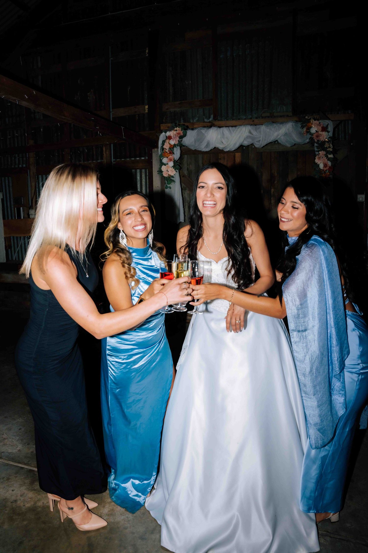 Bride in white ballgown toasts with three bridesmaids in blue and black dresses at barn wedding reception.