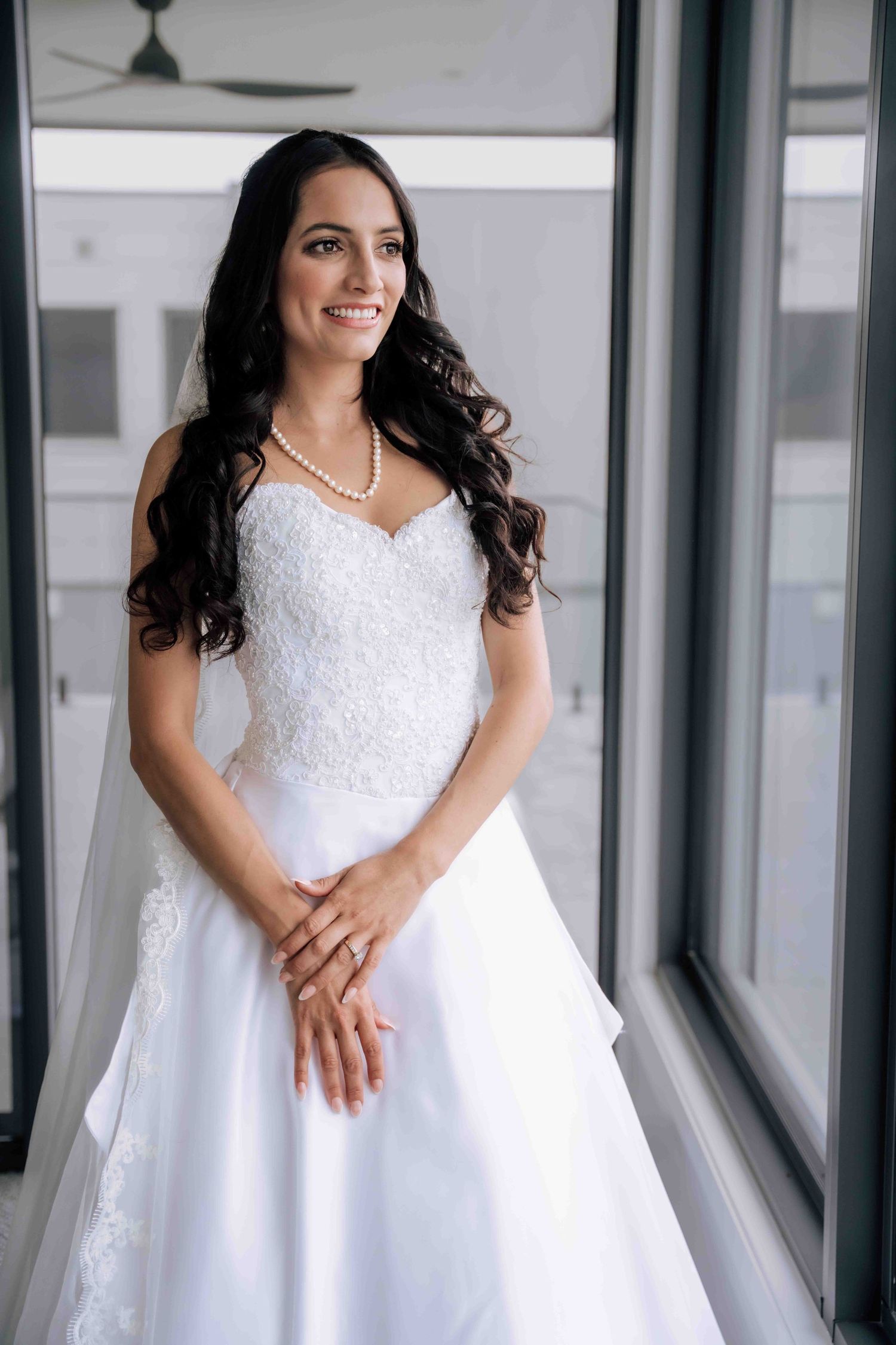 Elegant bride in strapless lace wedding gown with pearl necklace and veil standing by large windows.