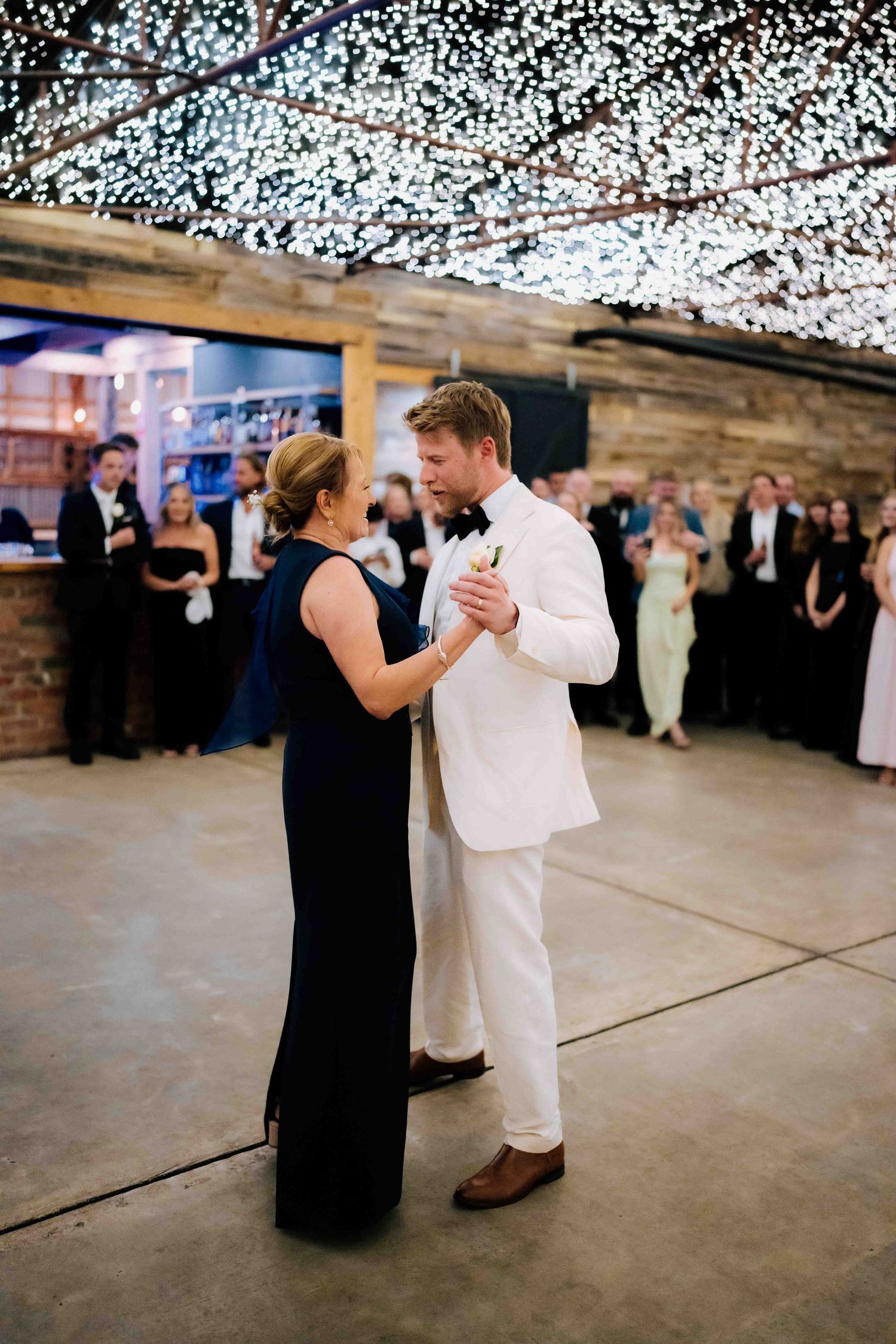 Groom in white jacket shares mother-son dance under fairy lights at wedding reception.