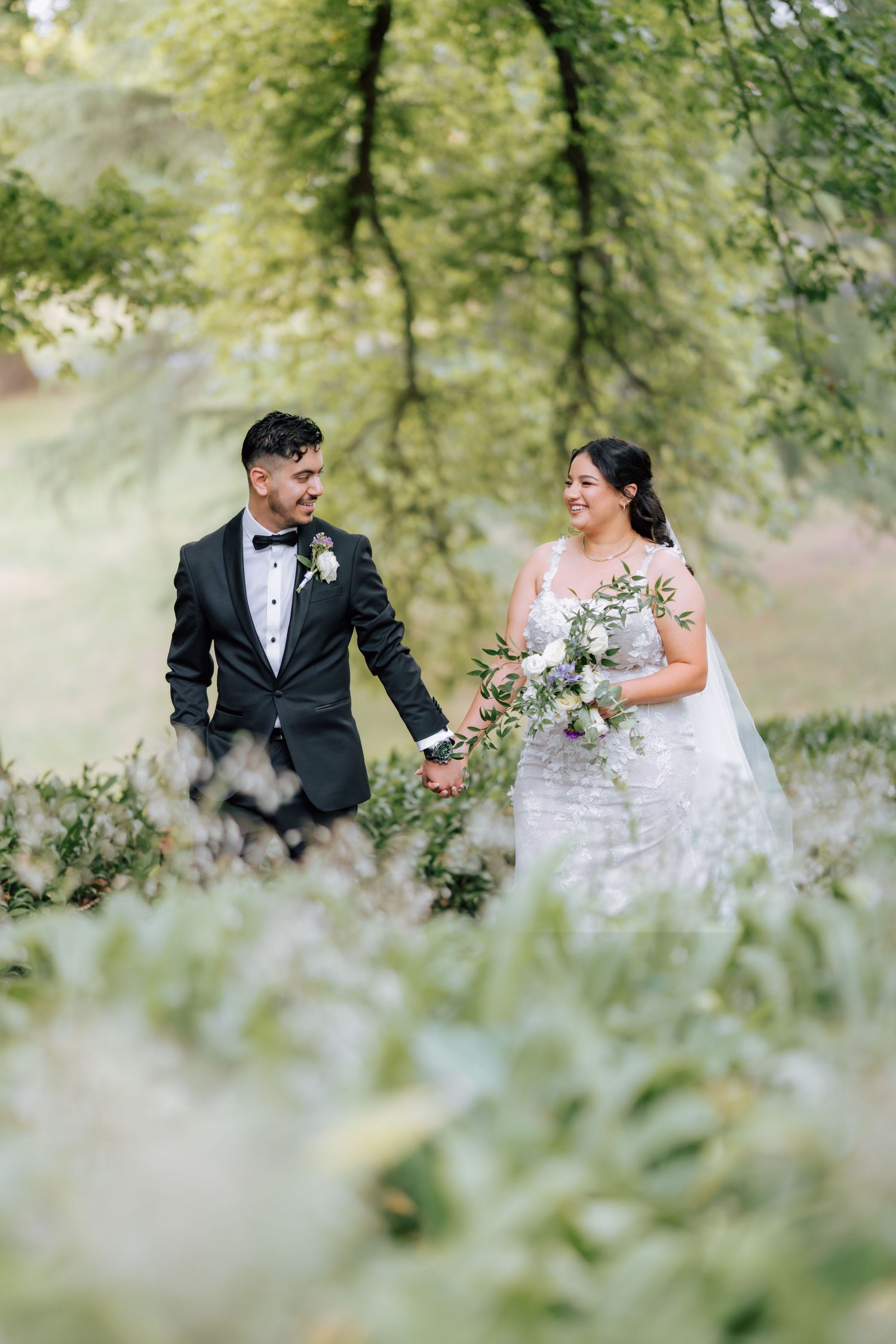 Bride and groom walk hand in hand through lush green garden, bride holding white floral bouquet.