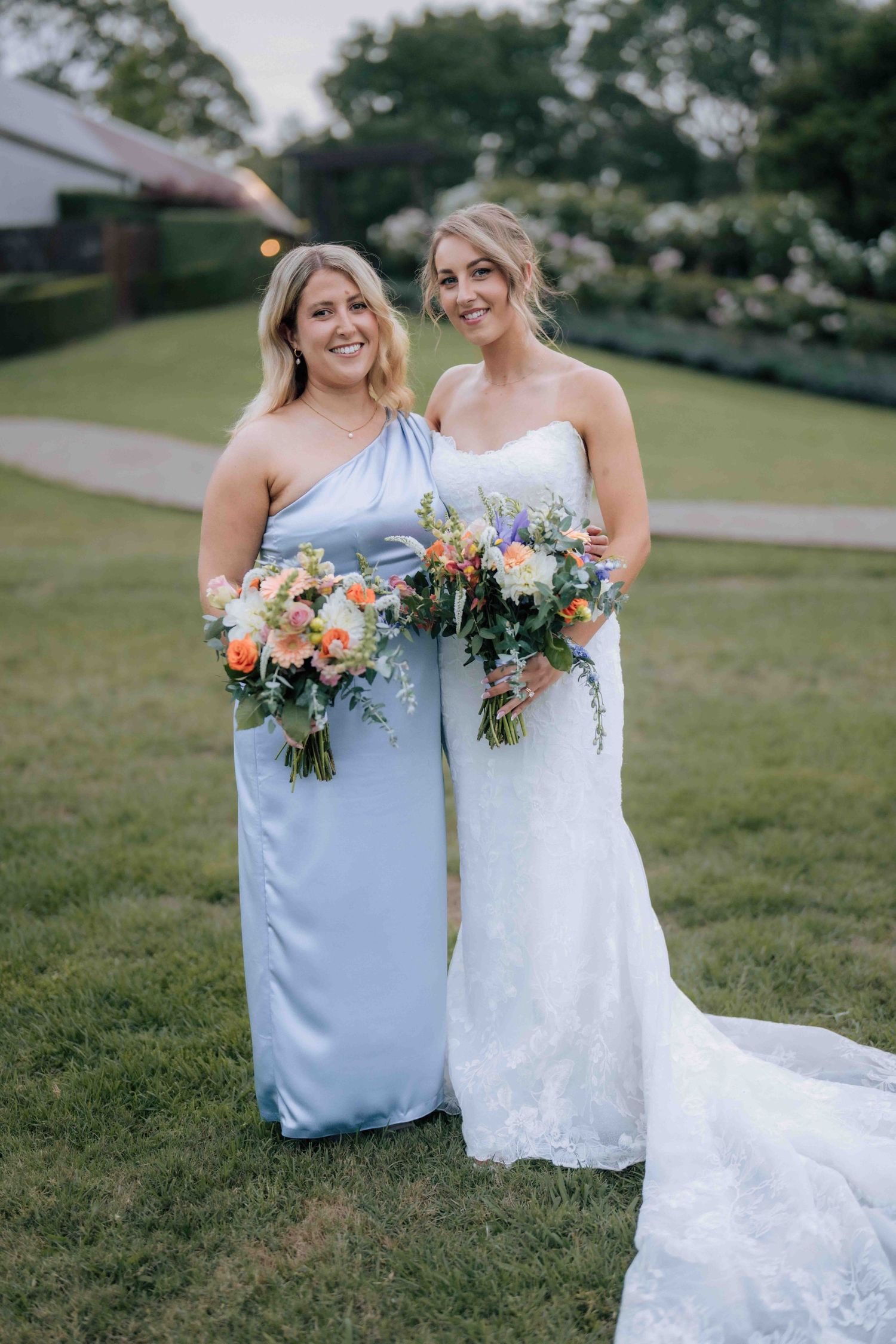 Blonde bride in strapless white lace gown poses with bridesmaid in one-shoulder light blue dress holding bouquets.