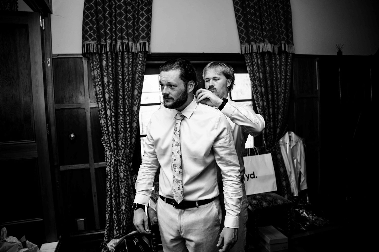 Black and white photo of two groomsmen getting ready, one adjusting the others tie indoors.
