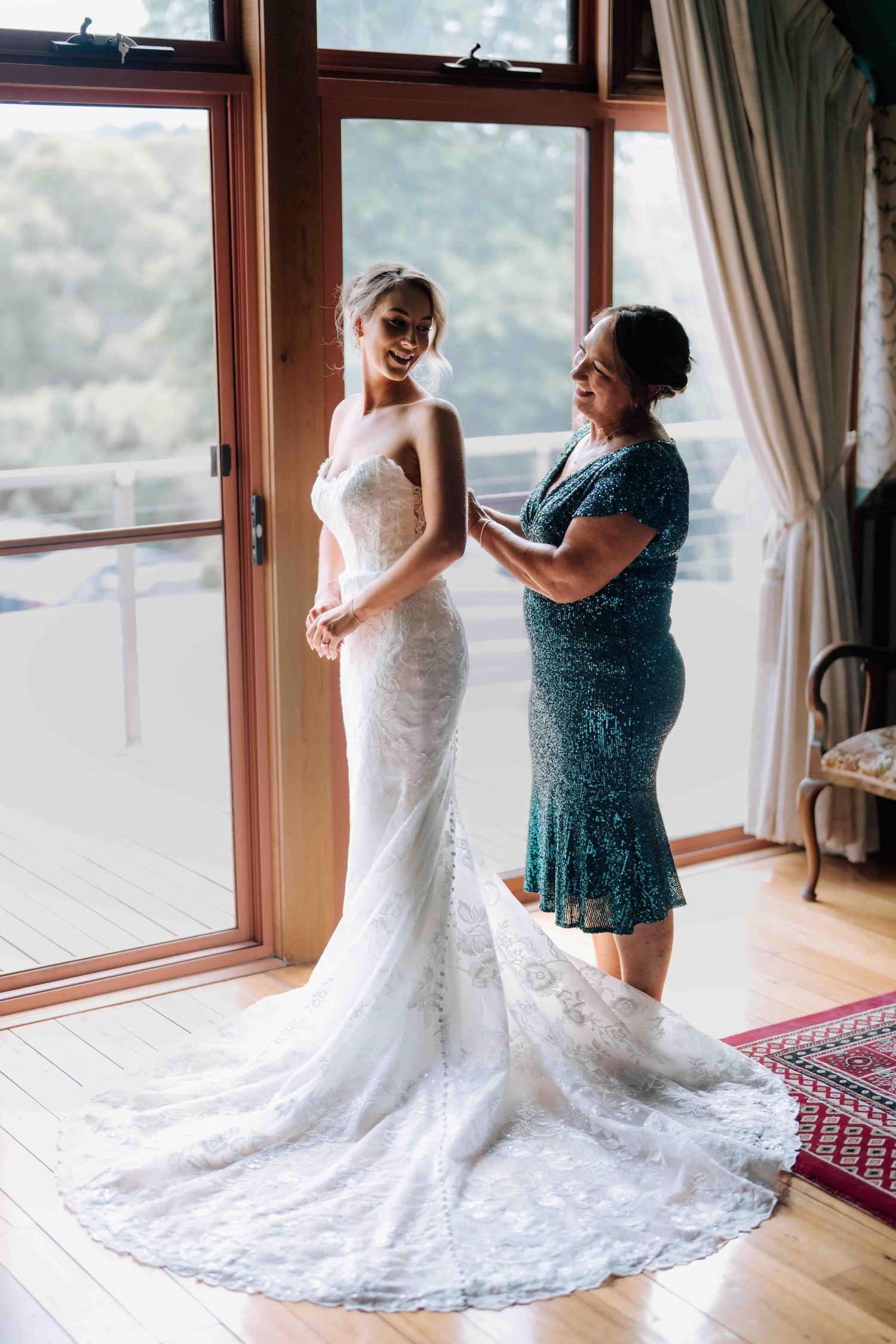 Mother helps bride button up her lace wedding gown near sunlit glass doors.