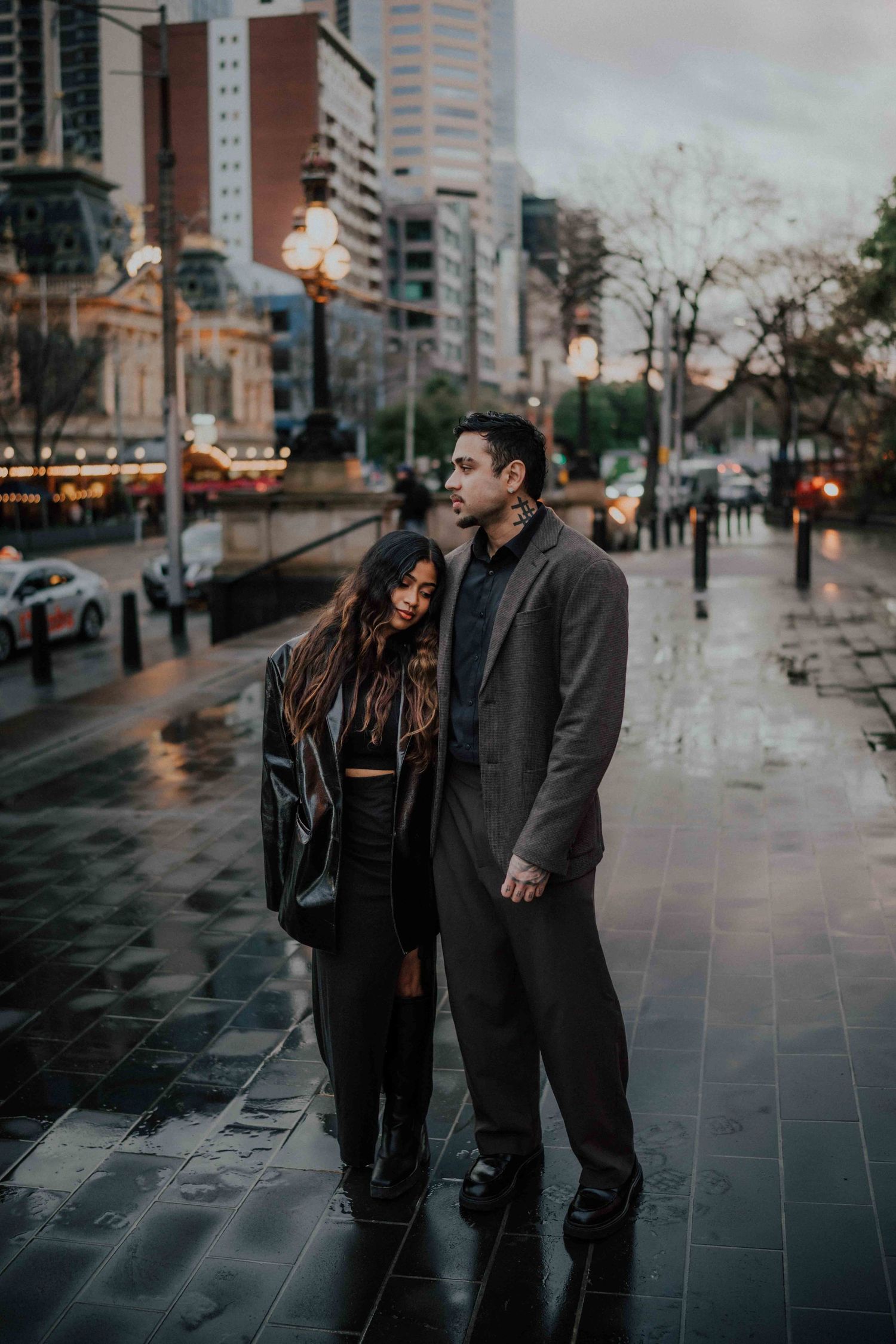 Stylish couple standing close together on wet city pavement during overcast evening in Melbourne.