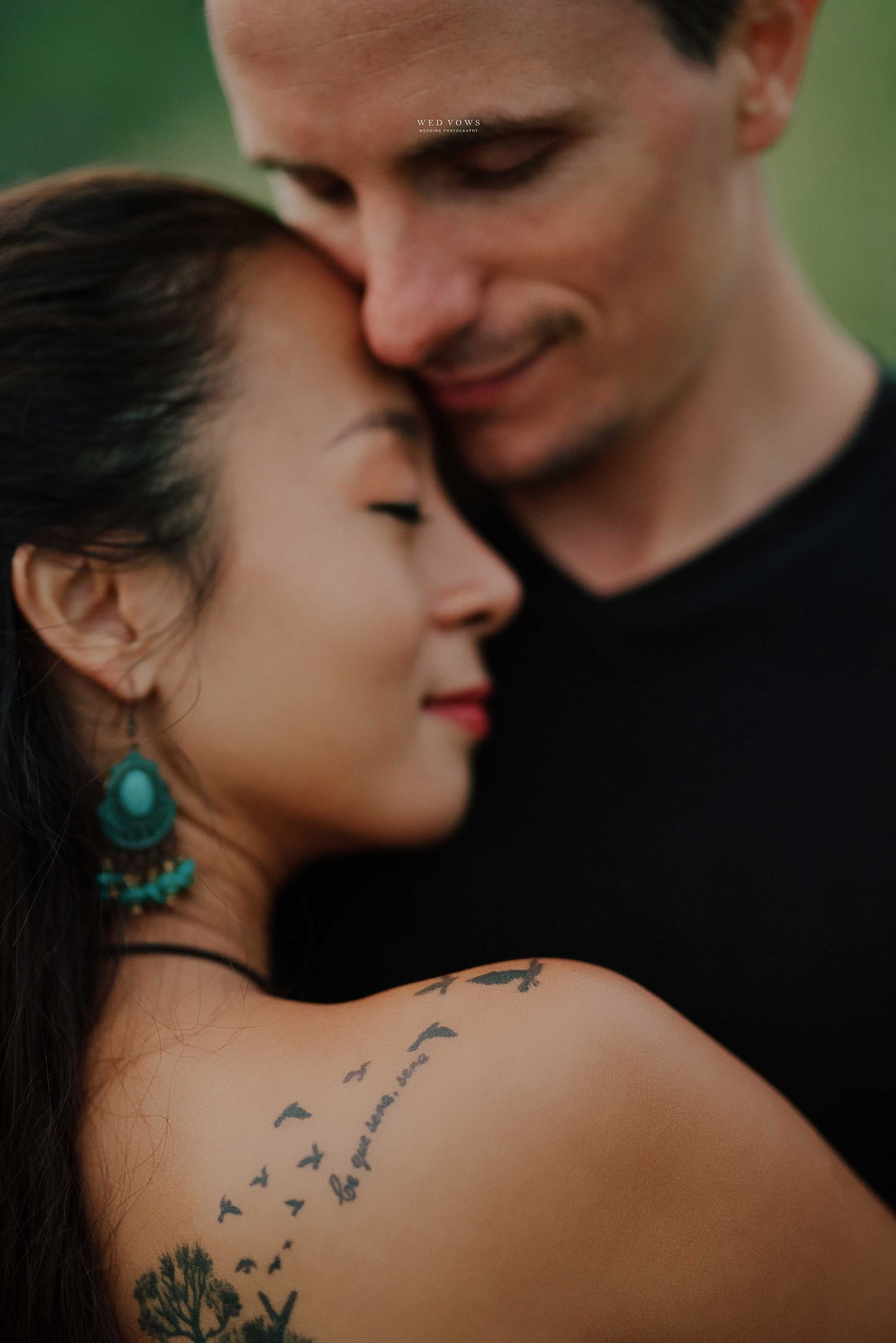 A woman with shoulder tattoo and teal earrings leans against a smiling man in a black shirt outdoors.