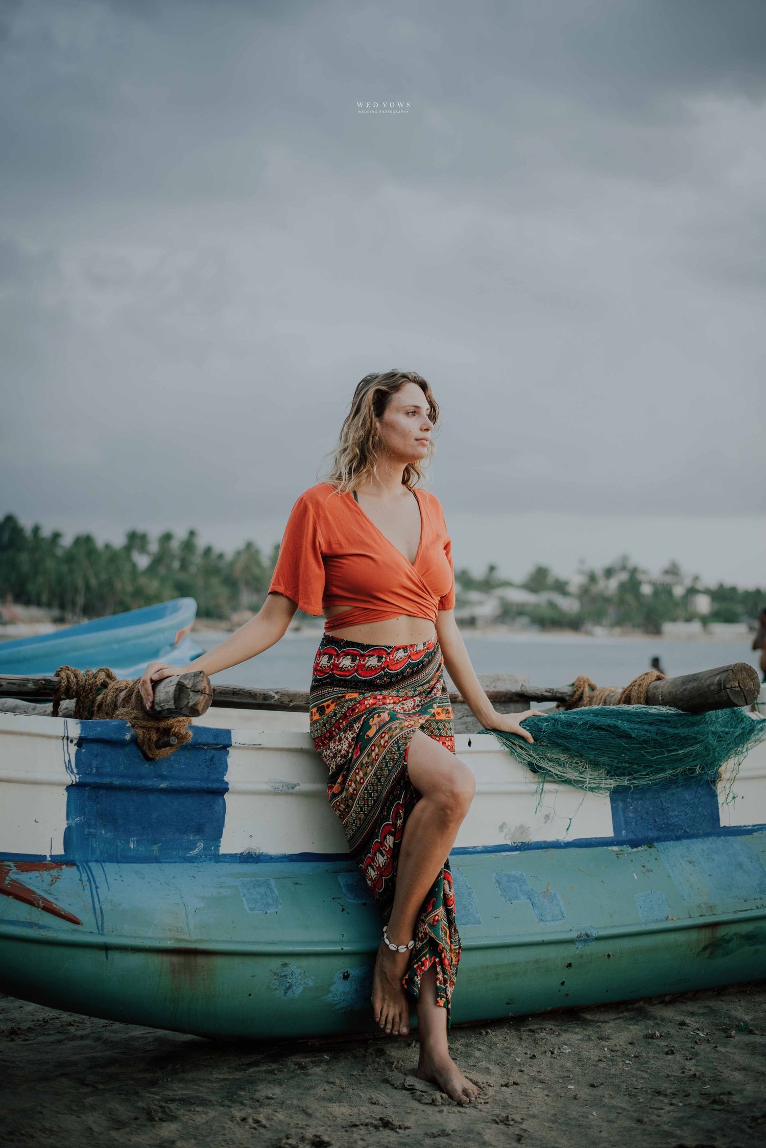 A woman in an orange wrap top and patterned skirt sits gracefully on a rustic fishing boat on the beach.