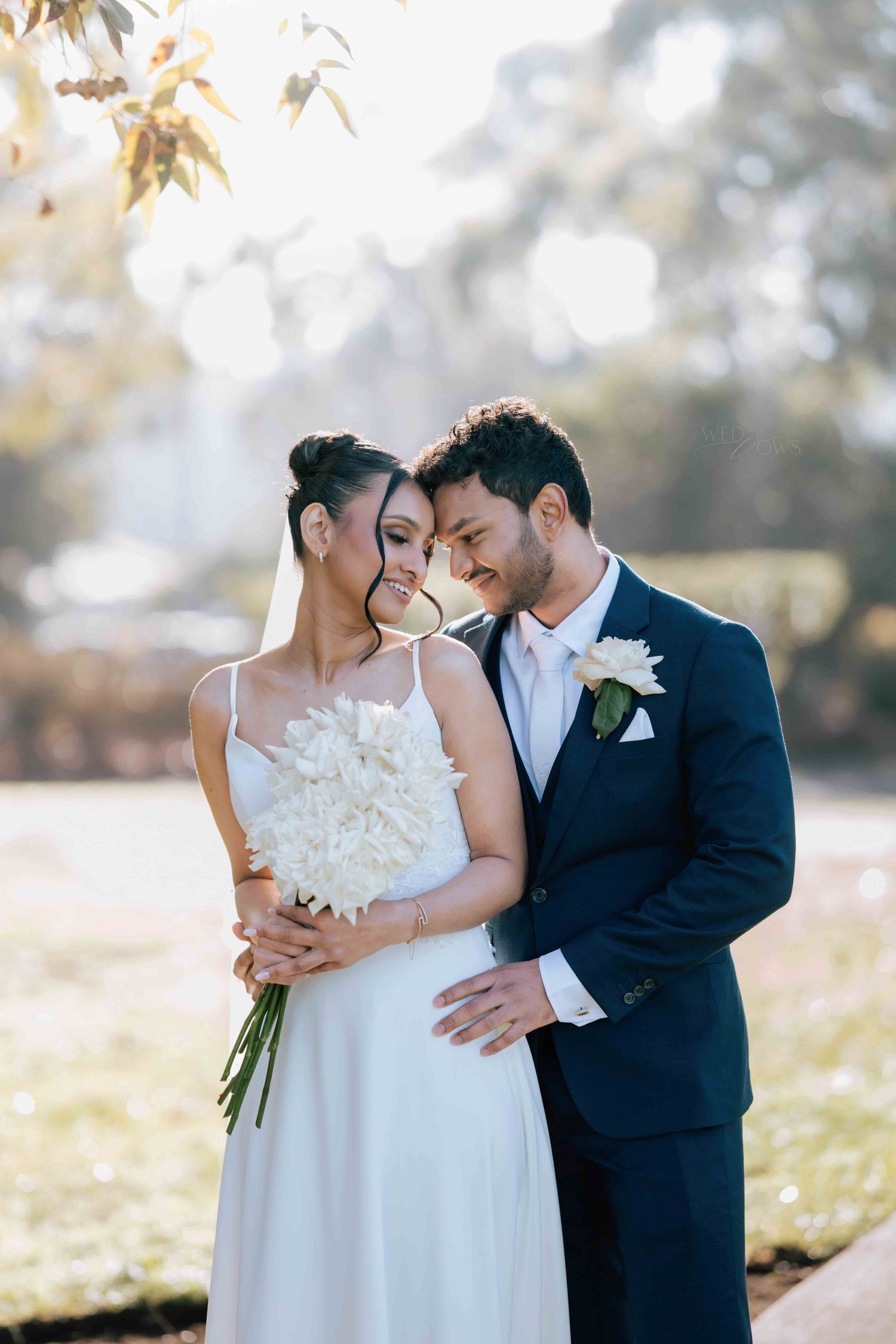 Bride and groom touch foreheads tenderly, bride holding white peony bouquet in golden sunlight.