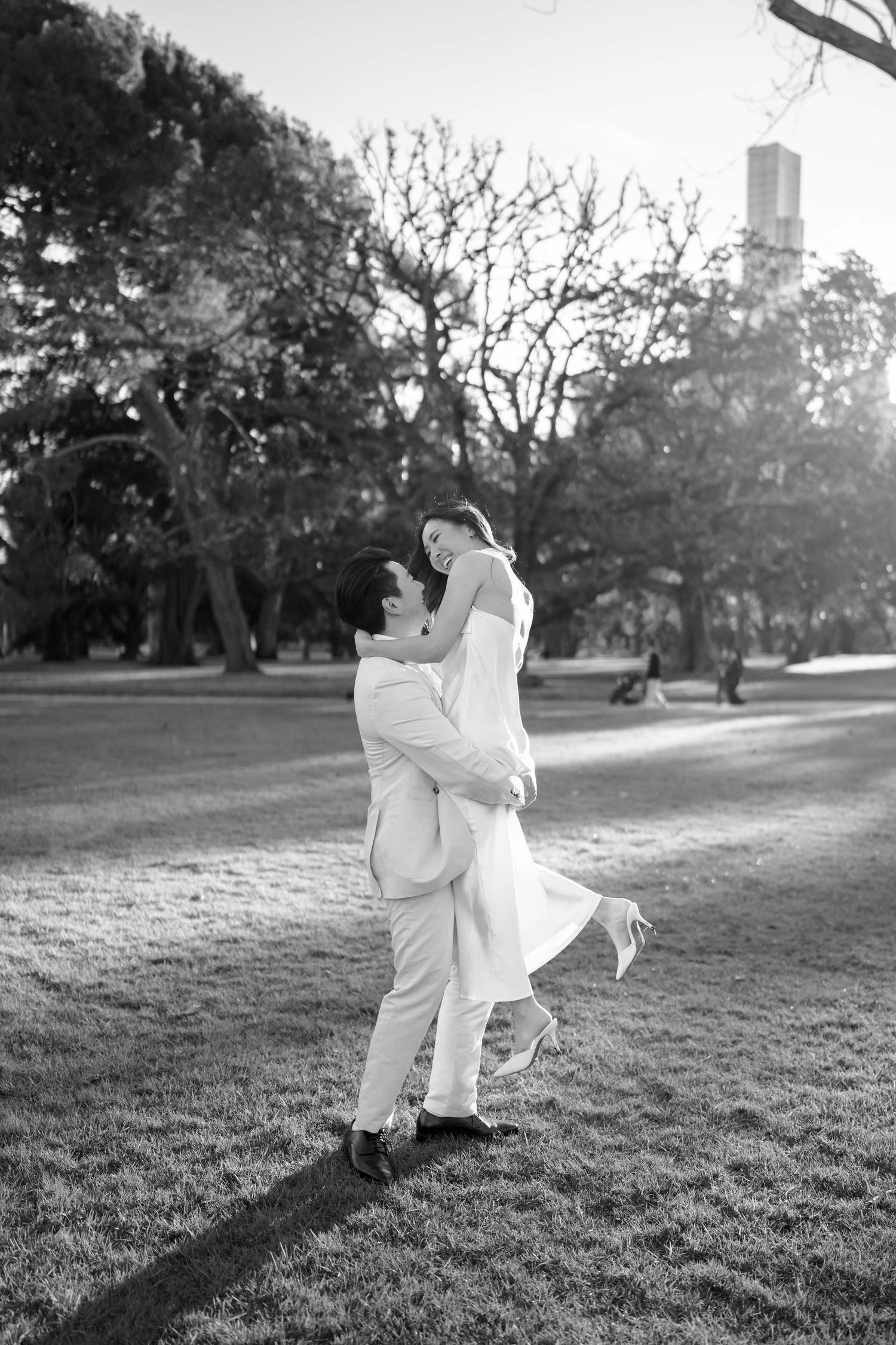 Black and white photo of a groom lifting his bride in a sunlit park, both dressed in white wedding attire.