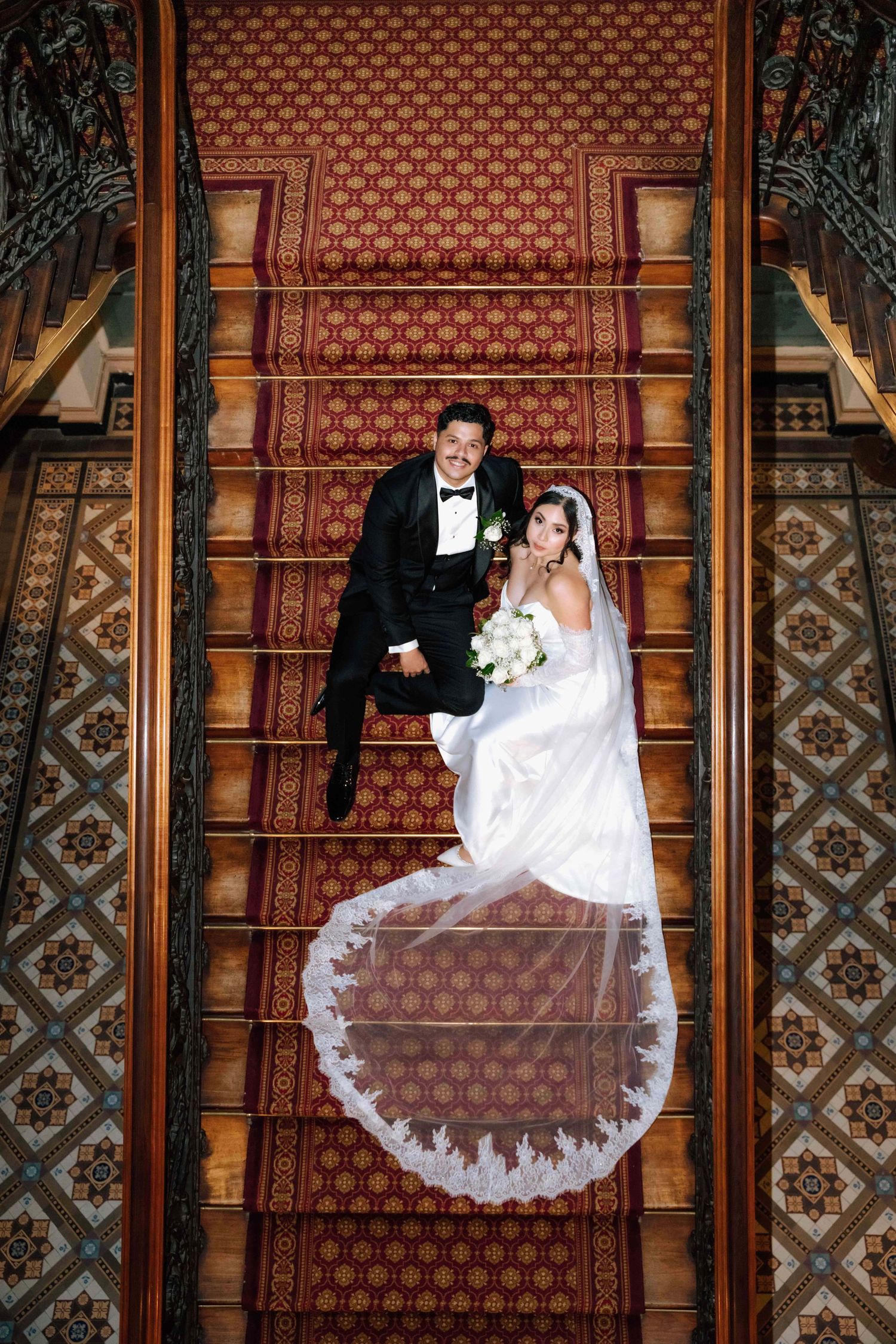 Bride and groom pose on ornate red carpeted staircase, bride's lace veil fans out beautifully in aerial wedding portrait.