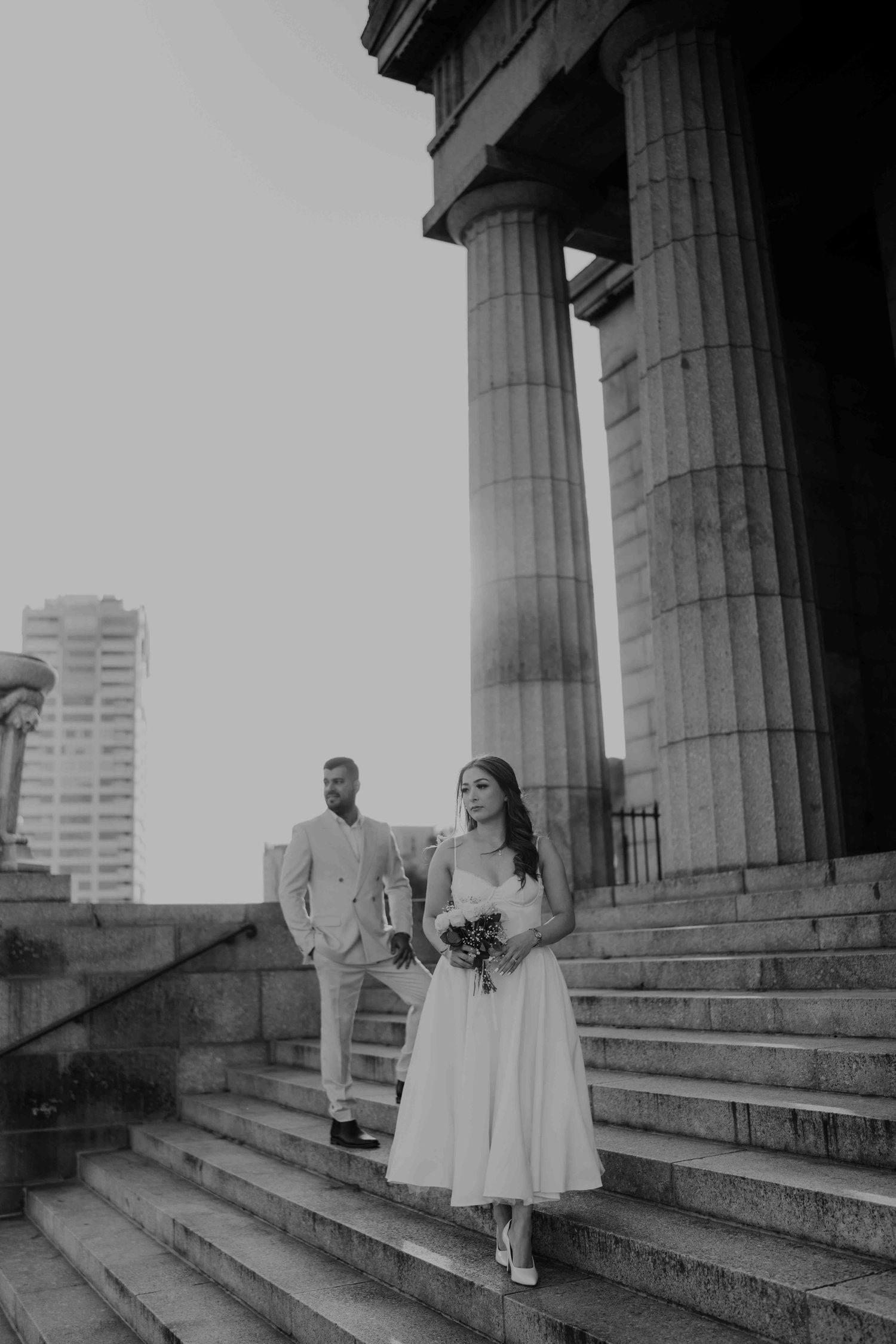 Black and white wedding portrait of bride descending stone steps with groom standing behind tall columns.