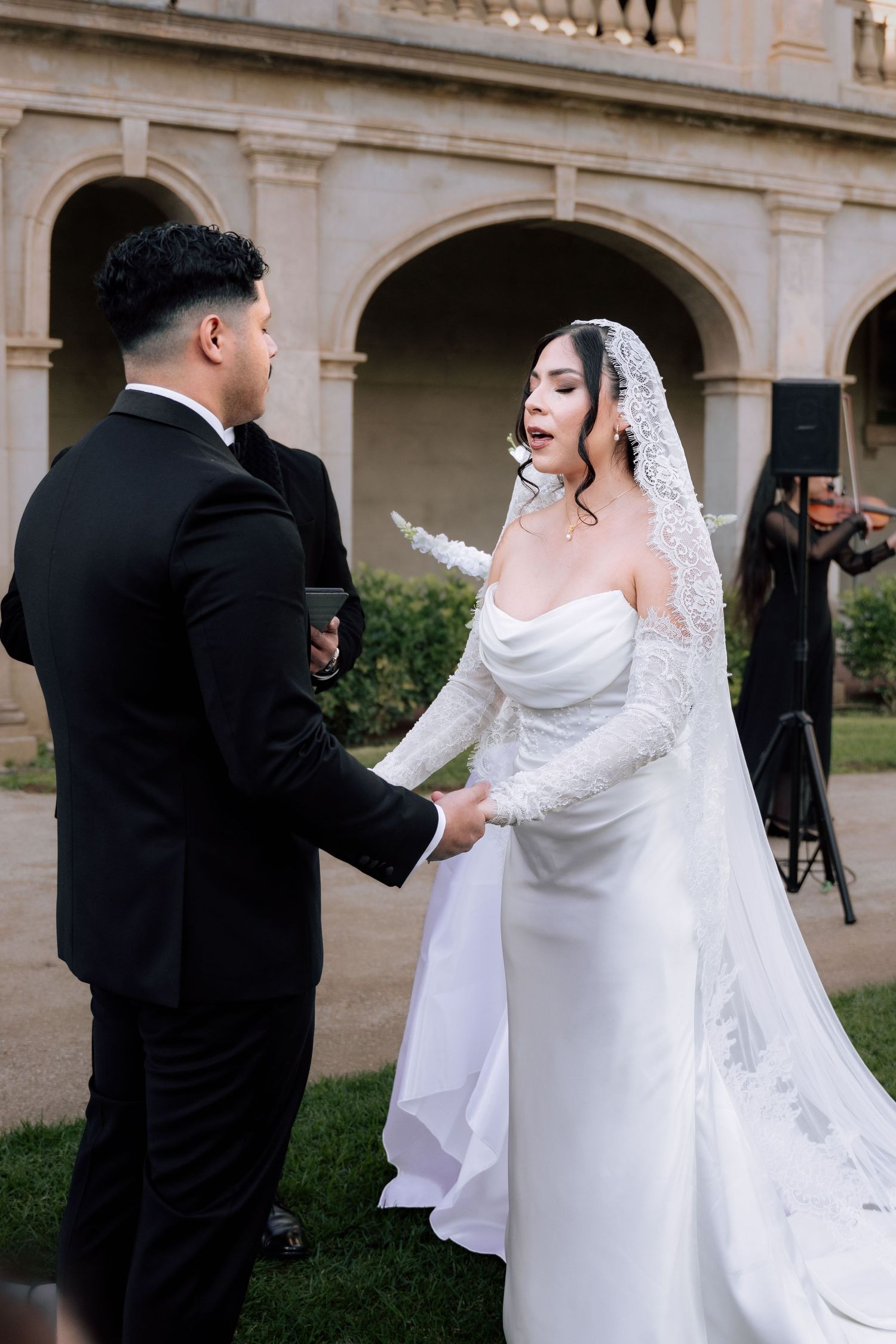 Bride in off-shoulder lace wedding gown and veil holding hands with groom during outdoor ceremony.