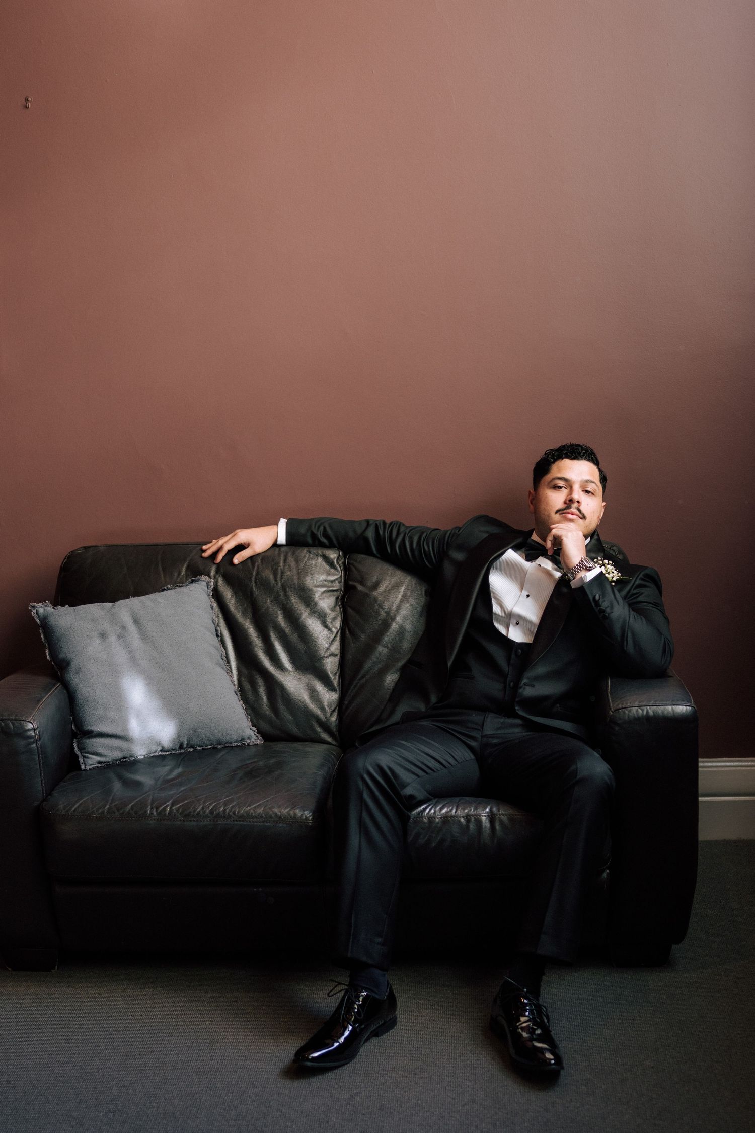 A well-dressed man in a black suit sits thoughtfully on a leather sofa against a mauve wall.