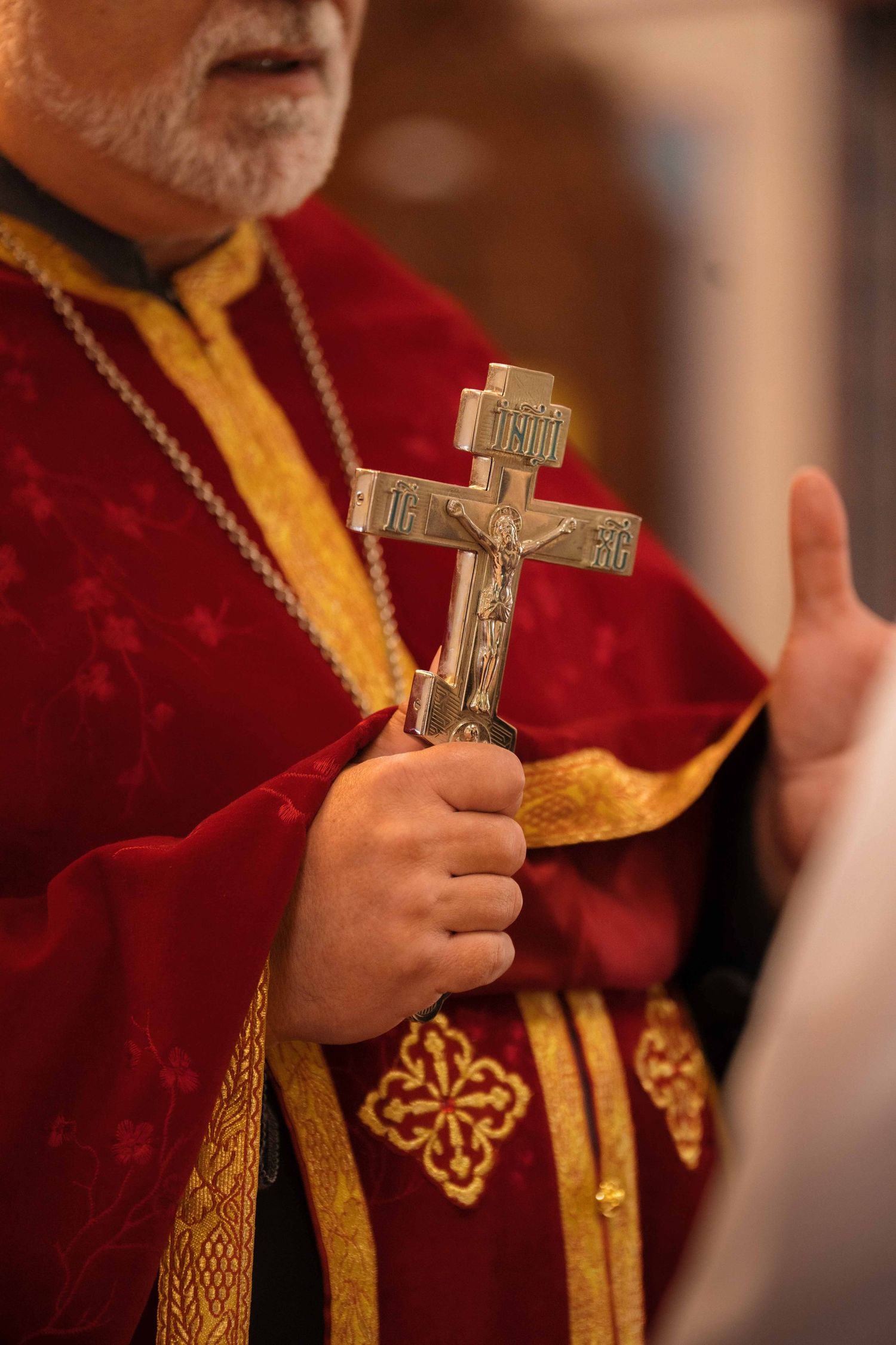 Orthodox priest in red and gold vestments holding a decorative silver crucifix during a wedding ceremony.