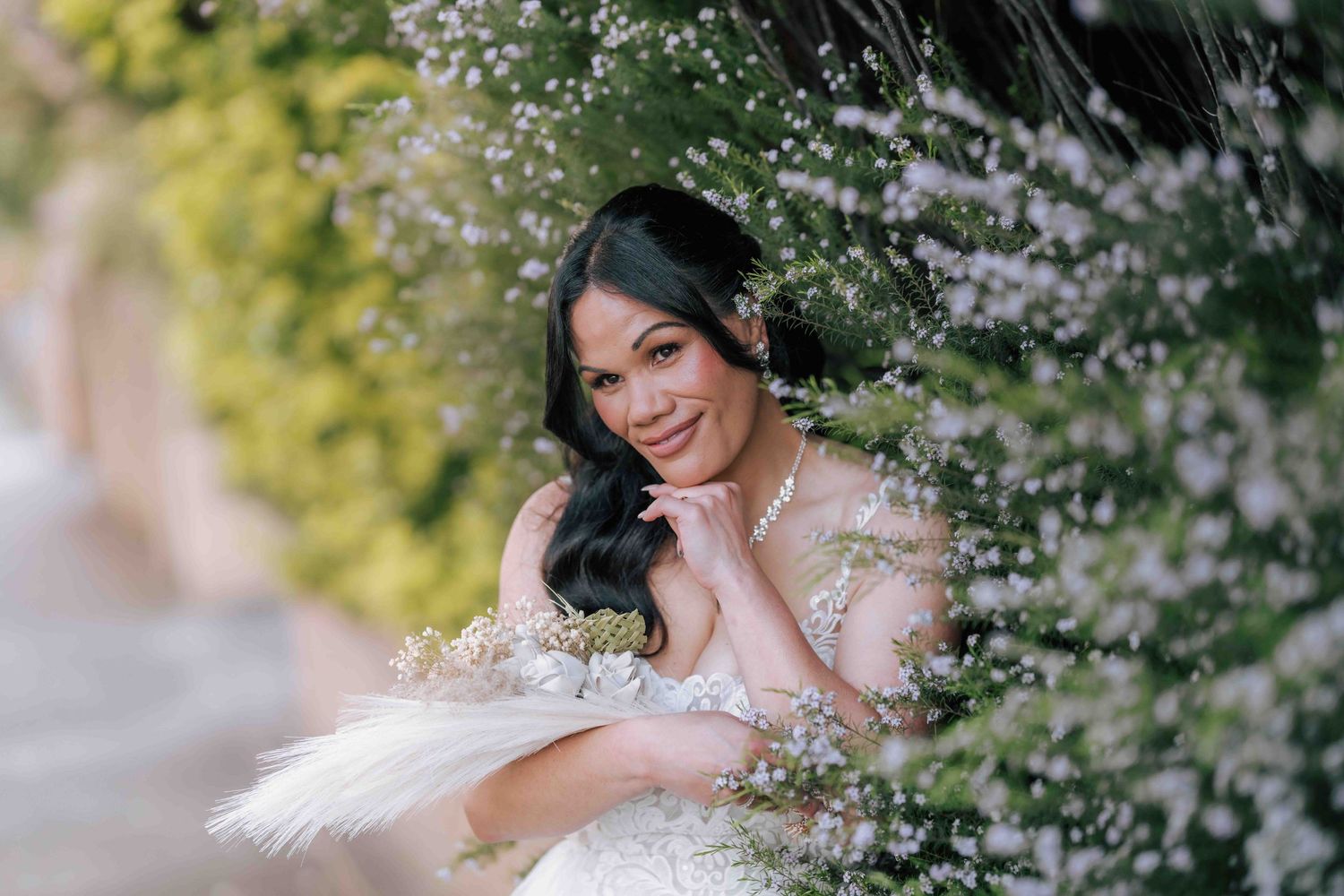 Radiant bride in lace gown posing among white flowering bushes with feather bouquet.