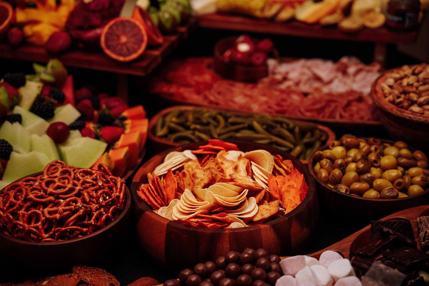 Close-up of grazing table with chips, pretzels, olives, dried fruits, and charcuterie in wooden bowls.