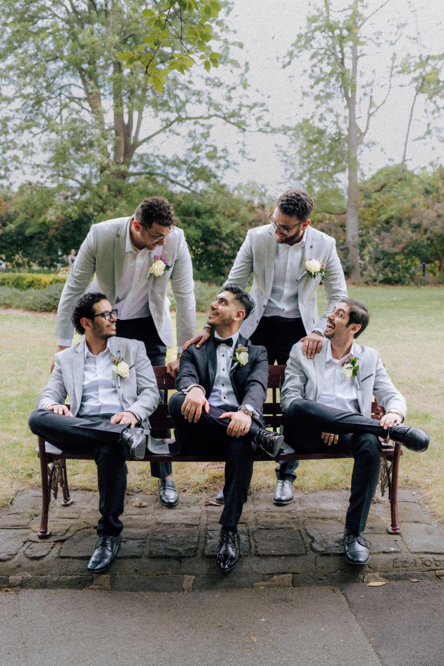 Groom and groomsmen in suits posing on bench outdoors at wedding.