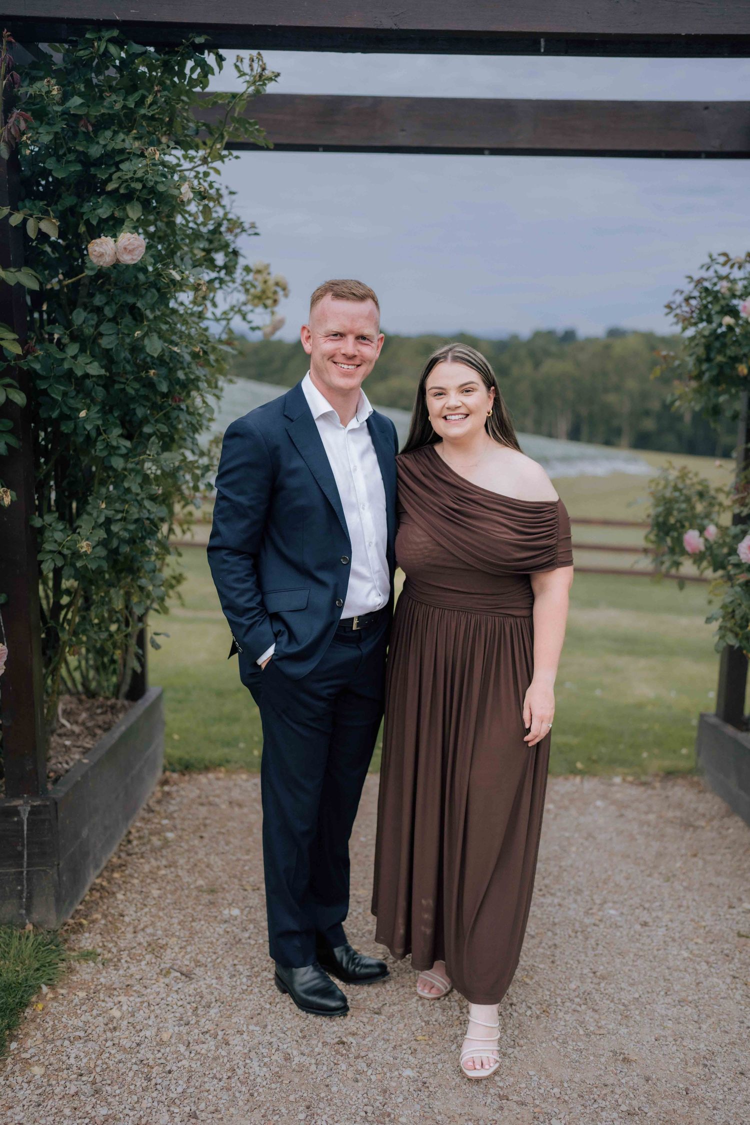 Couple in formal attire pose under a wooden pergola with scenic countryside views.