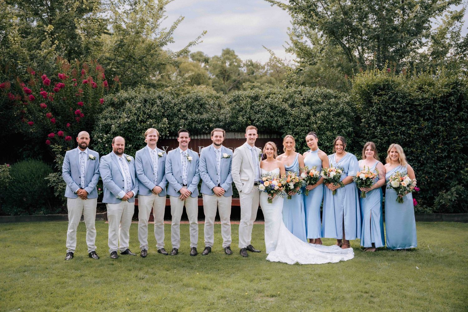 Wedding party in blue and cream attire poses in a line on green lawn with lush hedges.