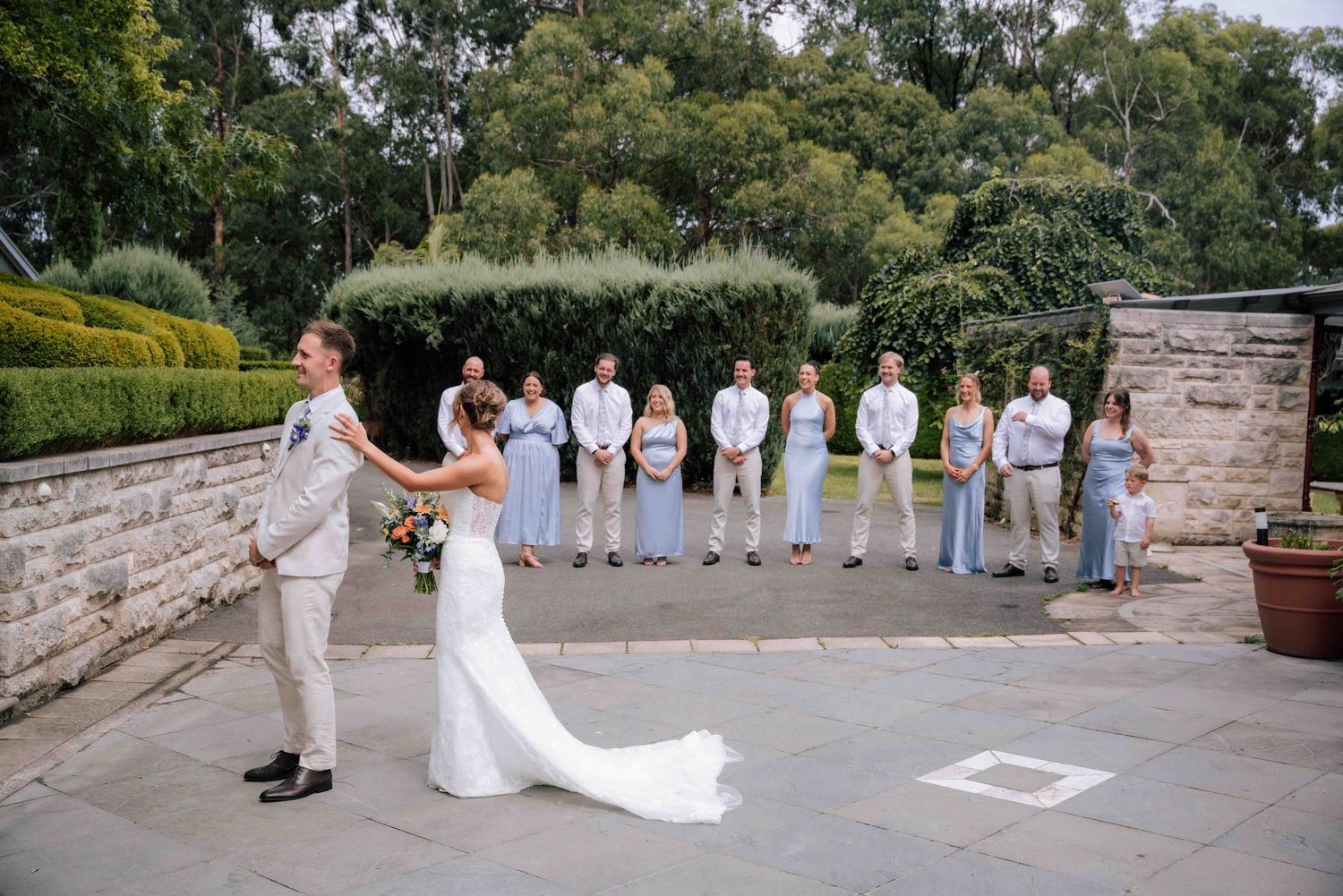 Bride and groom embrace during emotional first look while wedding party stands witness outdoors.