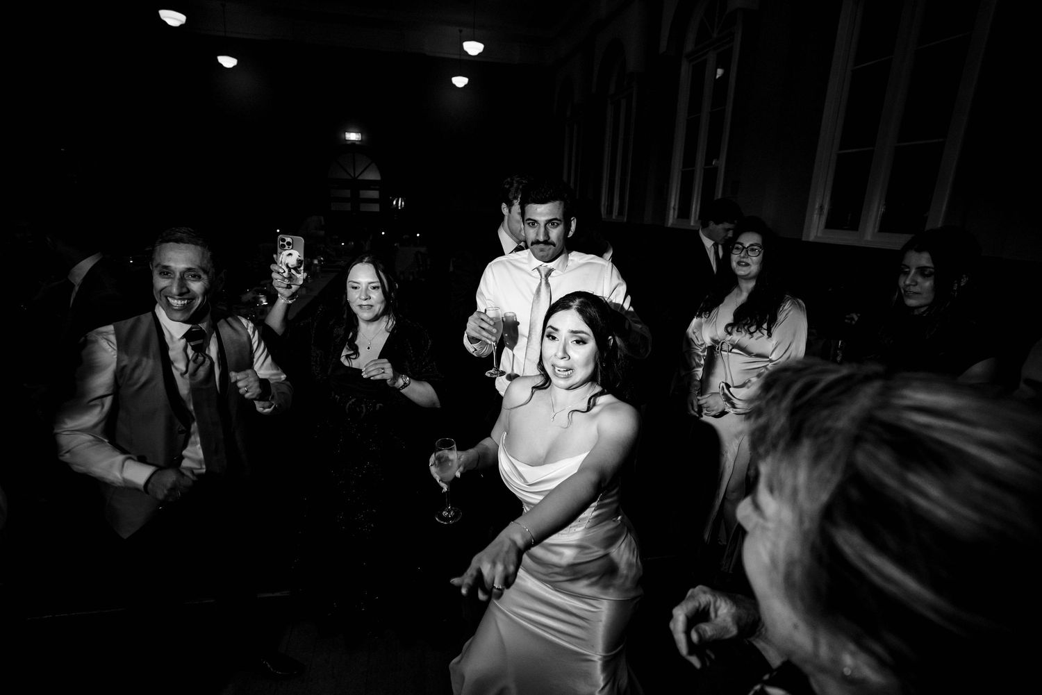 Bride dances joyfully with wedding guests in black and white photo on dimly lit reception dance floor.