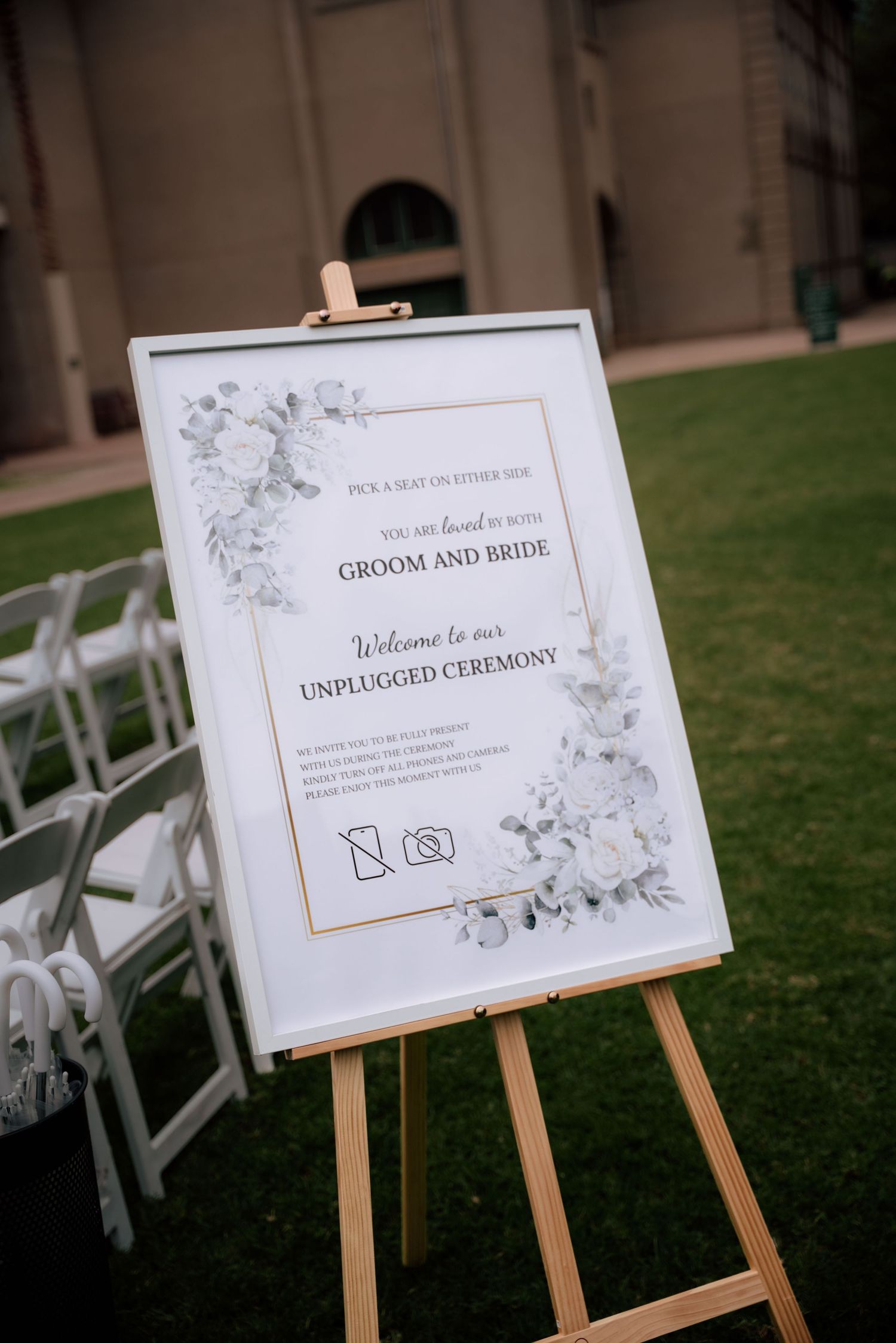 Elegant white floral wedding sign on wooden easel welcoming guests to an unplugged outdoor ceremony.