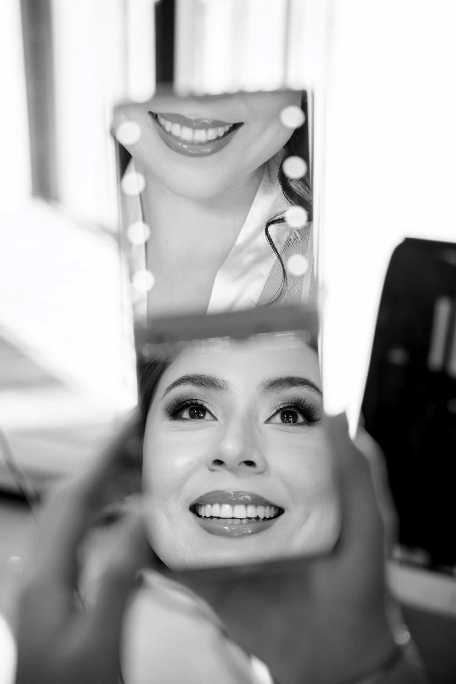 Black and white photo of a smiling bride checking her makeup in a handheld mirror.