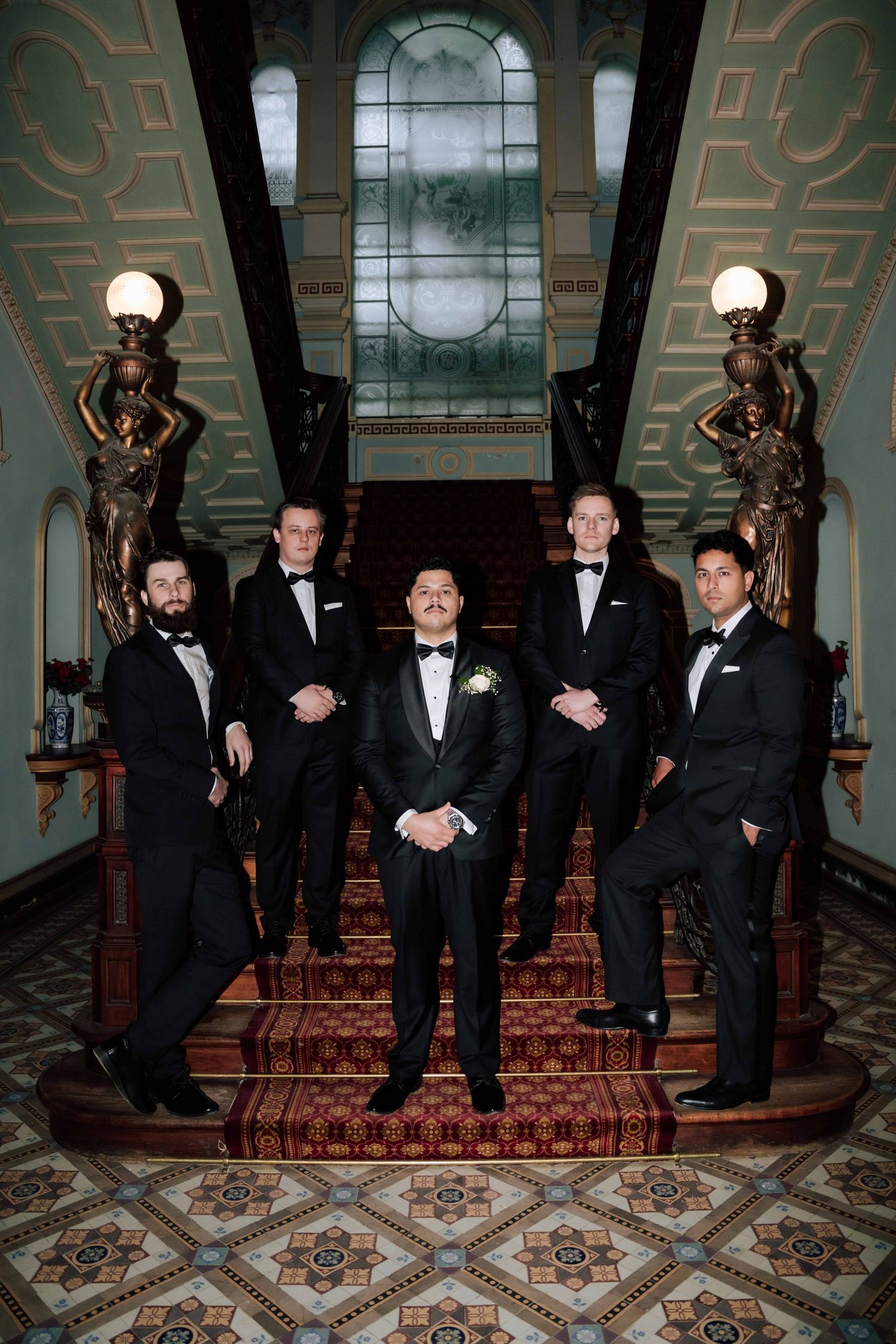 Groom and groomsmen in black tuxedos posing on ornate staircase in elegant venue.