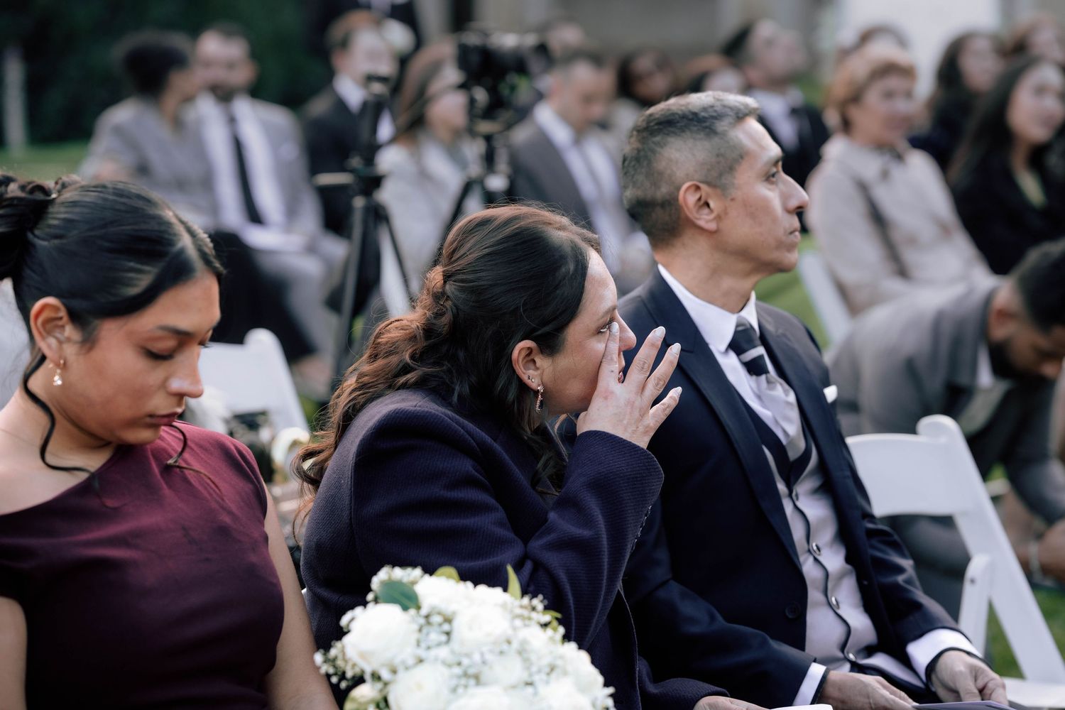 Emotional wedding guests seated outdoors, woman wiping tears while holding white flower bouquet.