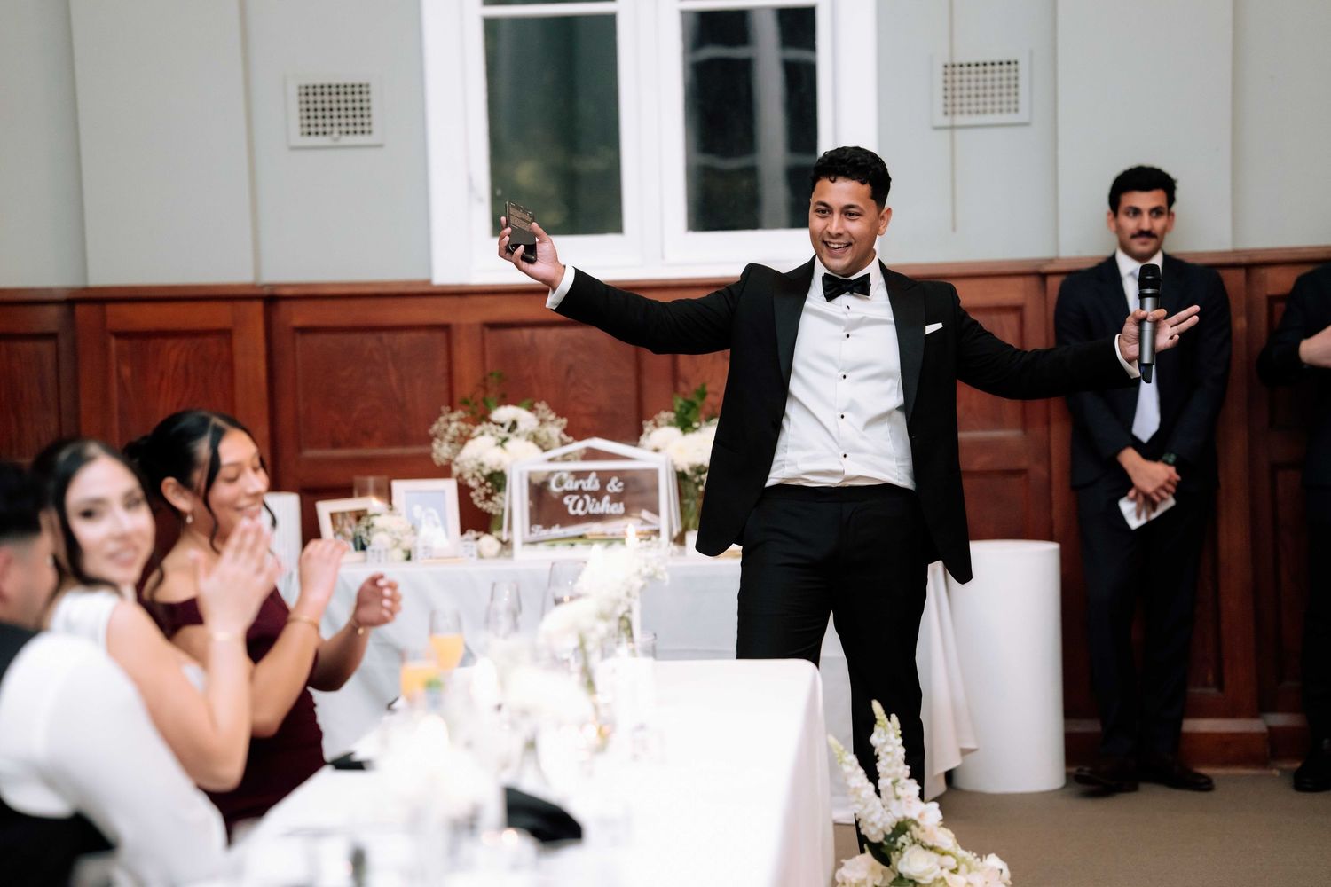 Groom in tuxedo raises glass giving energetic wedding speech at elegantly decorated reception table.