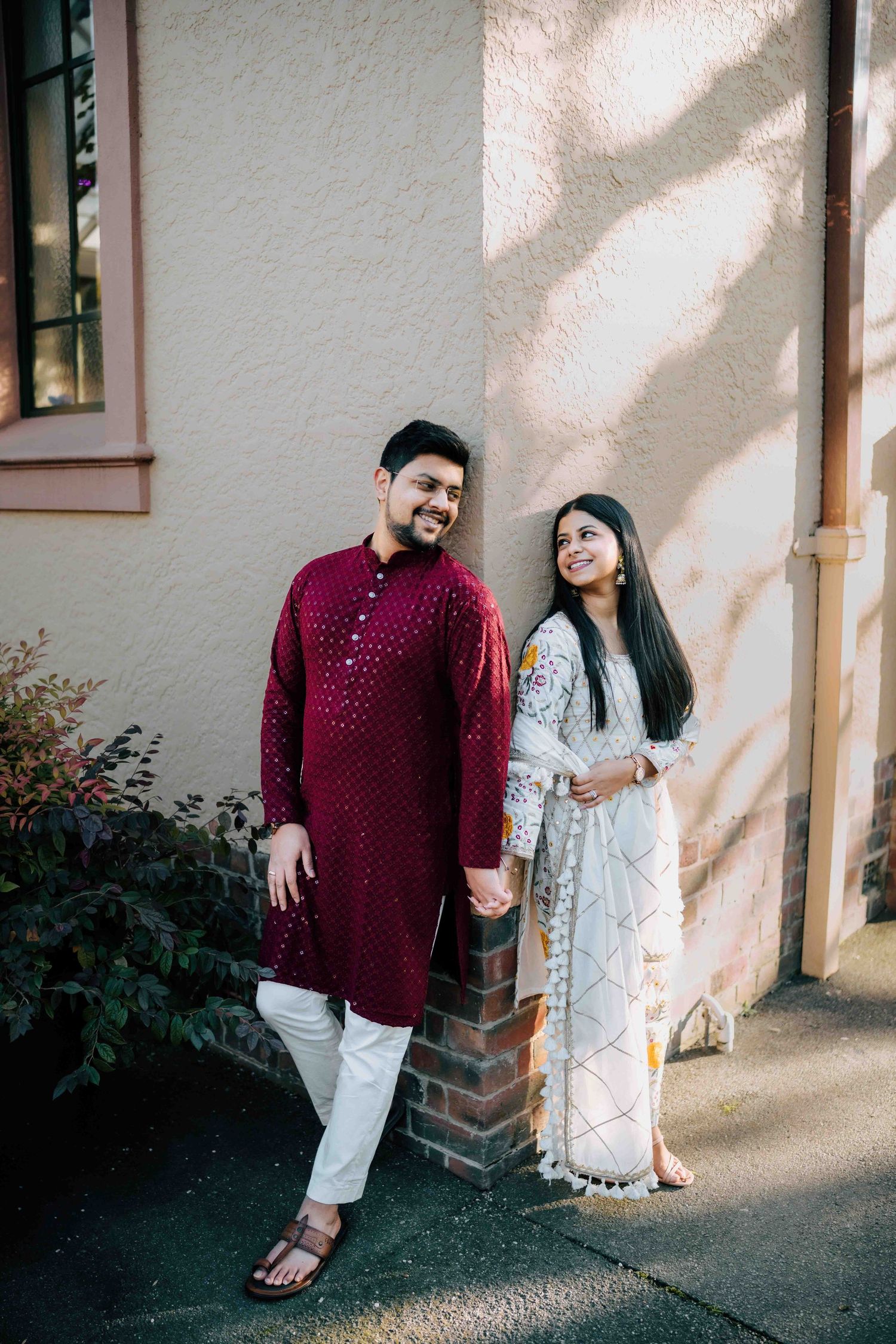 Smiling South Asian couple in traditional kurta outfits posing against cream building wall outdoors.