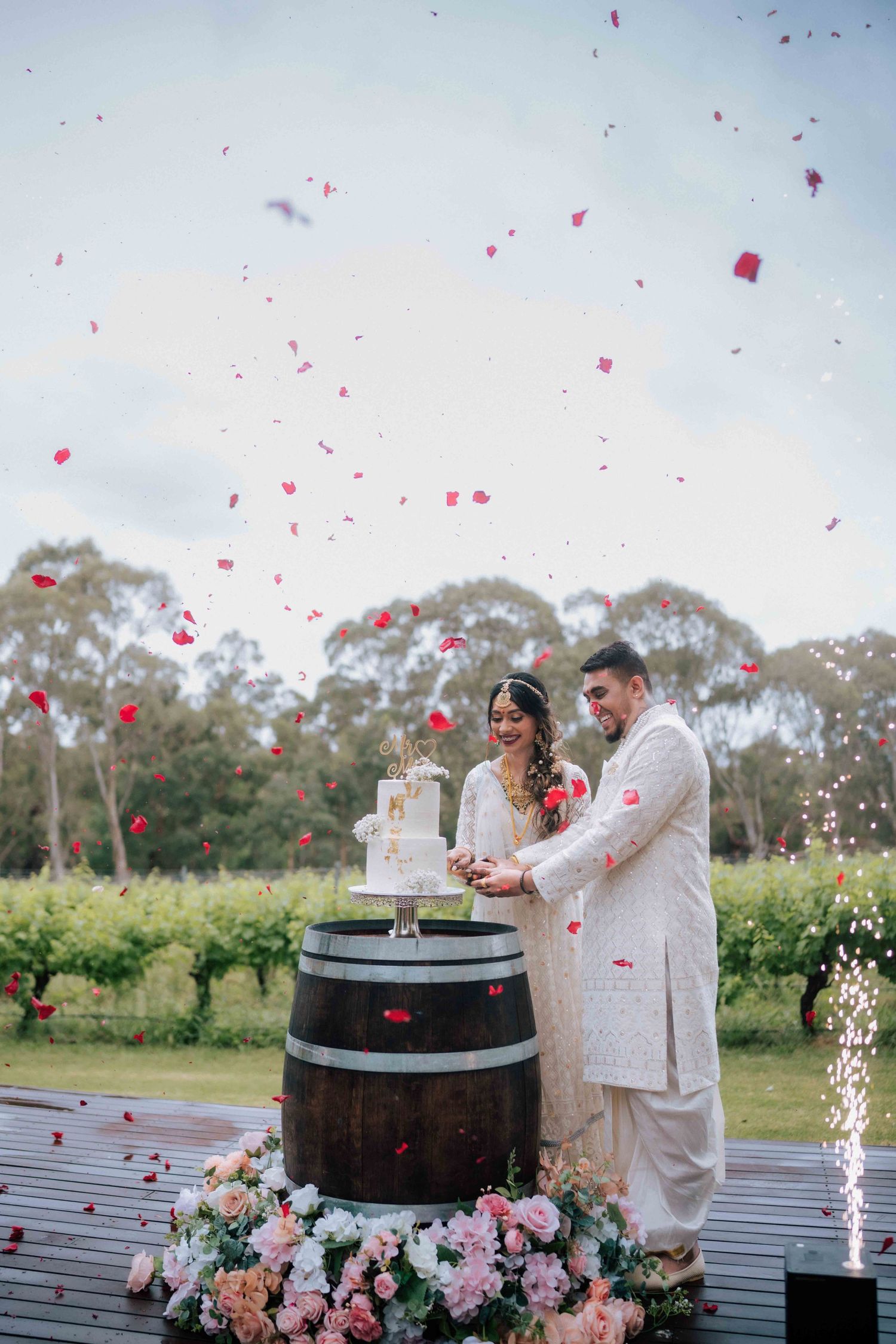 Indian couple cutting wedding cake on barrel with rose petals falling and fireworks at vineyard wedding.