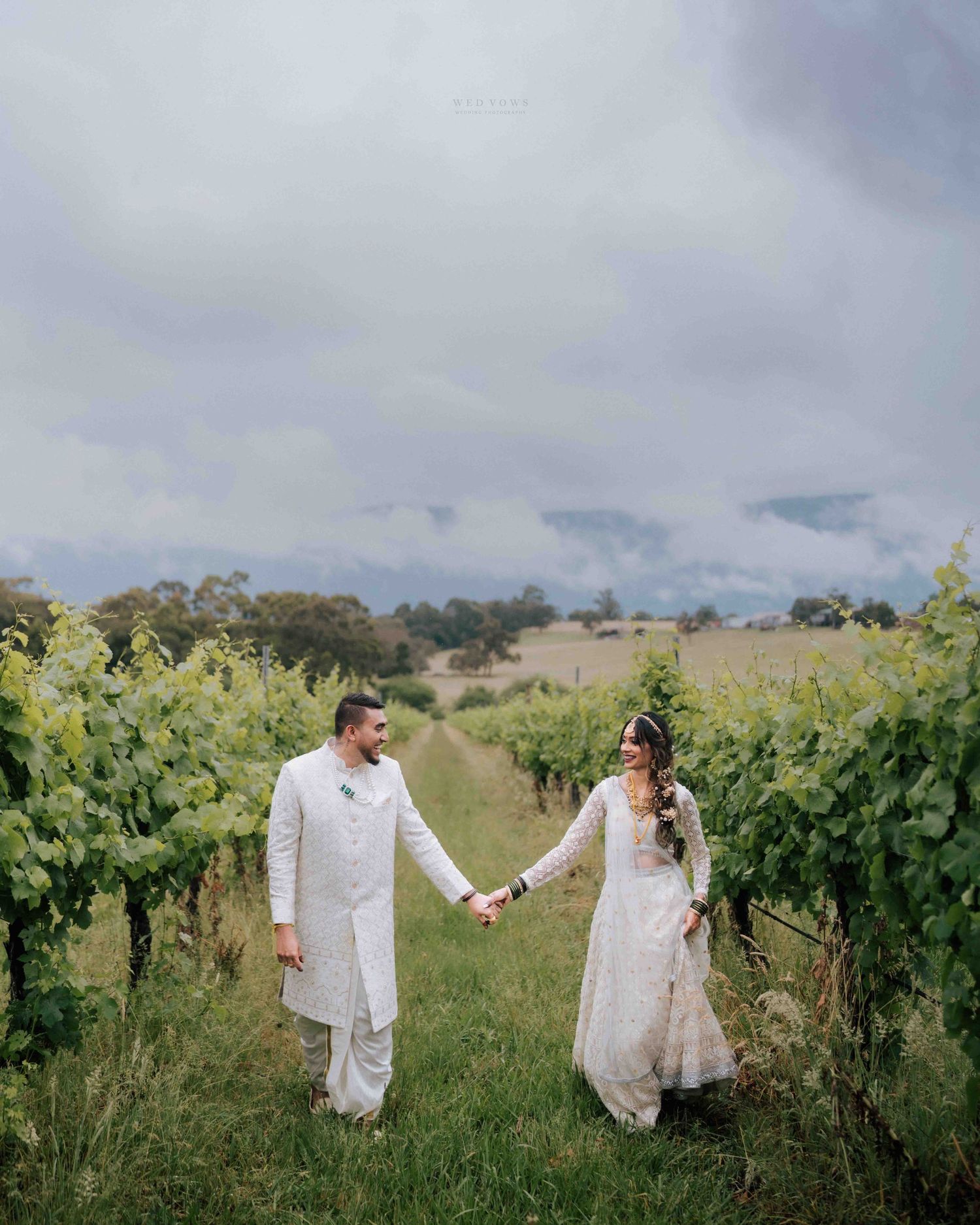 Indian wedding couple in white traditional attire hold hands walking through a scenic vineyard.