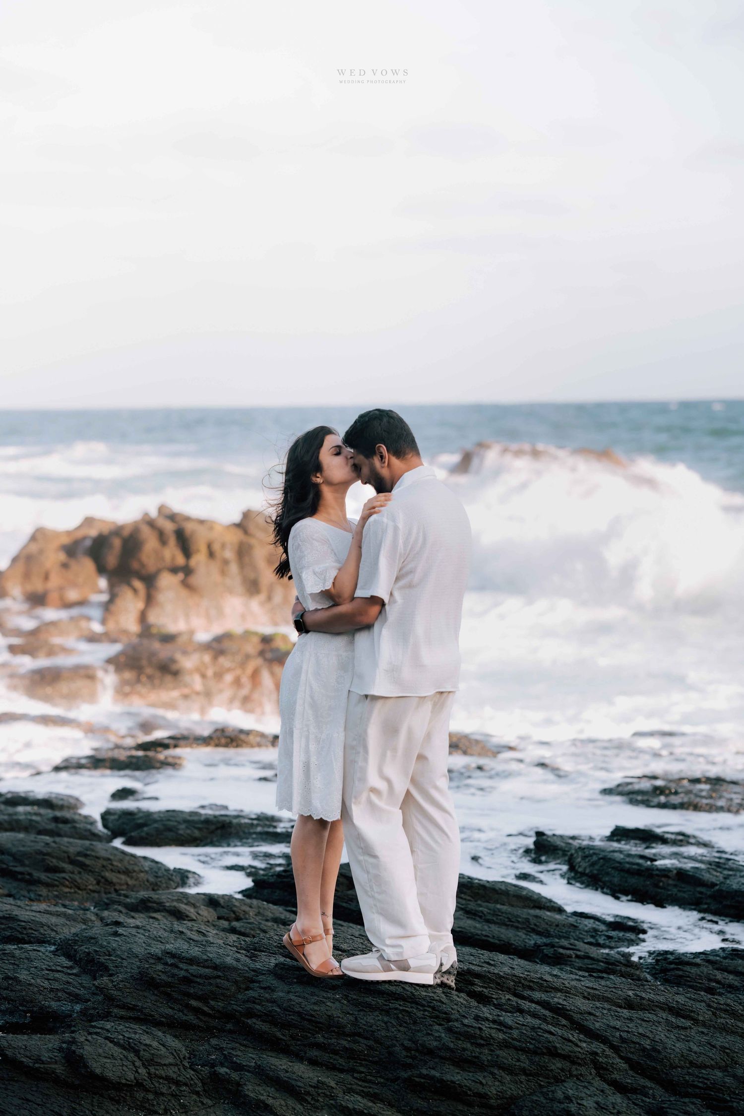 Couple in white sharing a romantic kiss on rocky shores with crashing waves behind.