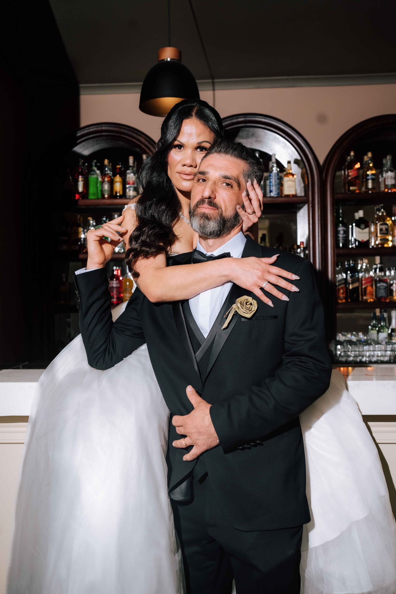 A bride in a white ball gown embraces her groom in a black tuxedo near an elegant bar setting.