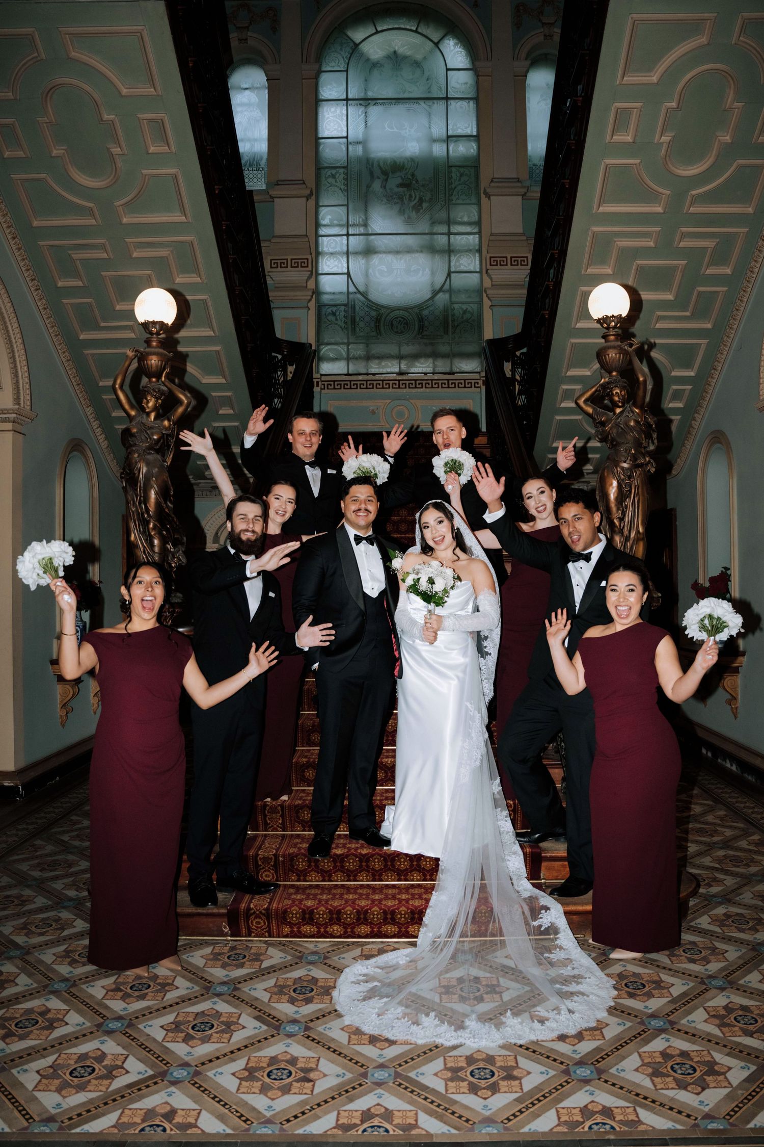 Wedding party celebrates on a grand staircase with ornate statues and stained glass window.