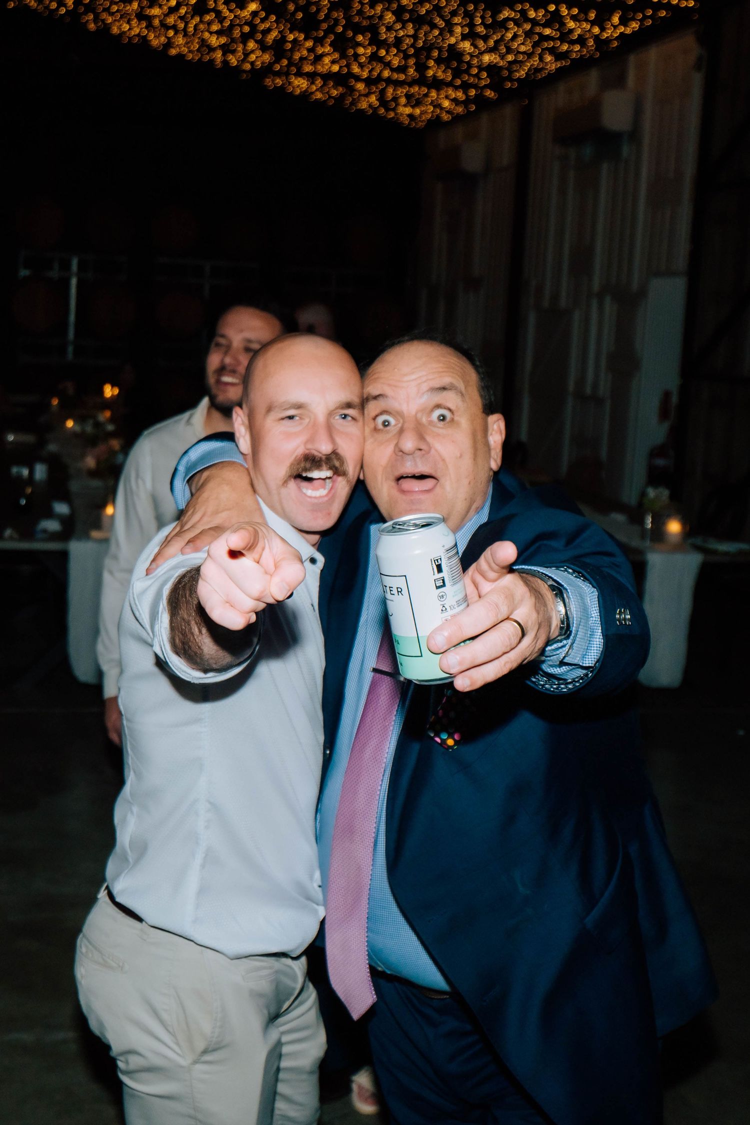 Two men smiling and pointing at camera during wedding reception, one holding a beer can.