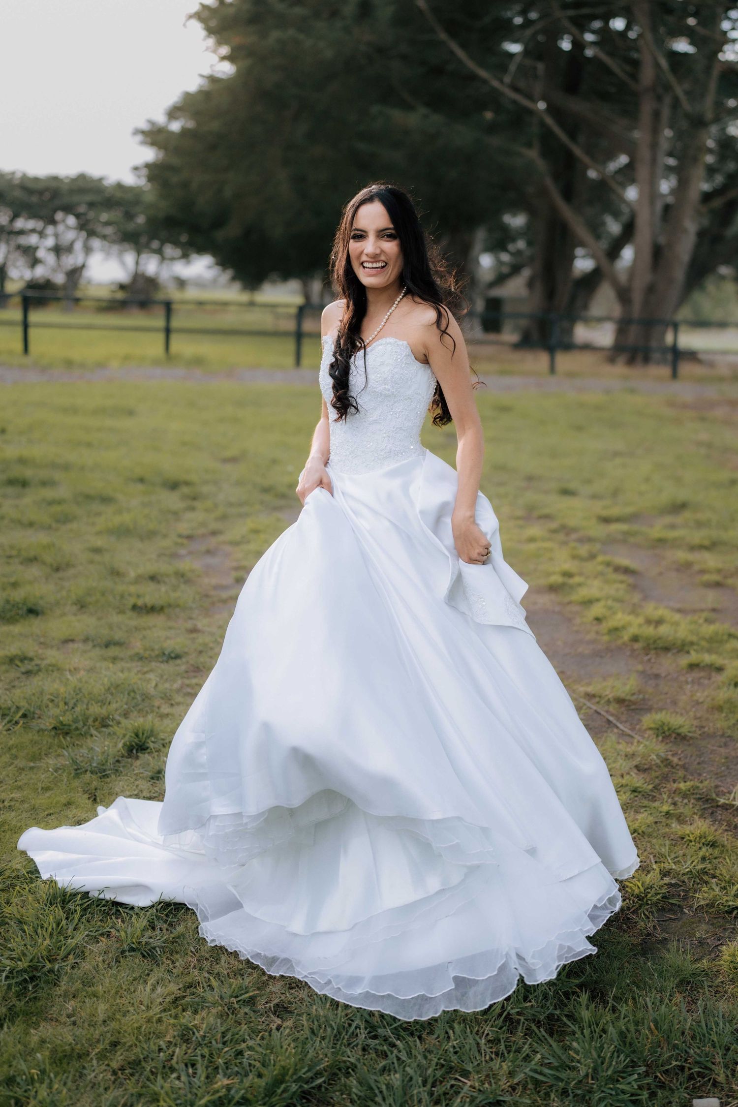 Smiling bride in a flowing white ball gown with a long train poses outdoors on green grass with trees behind her.