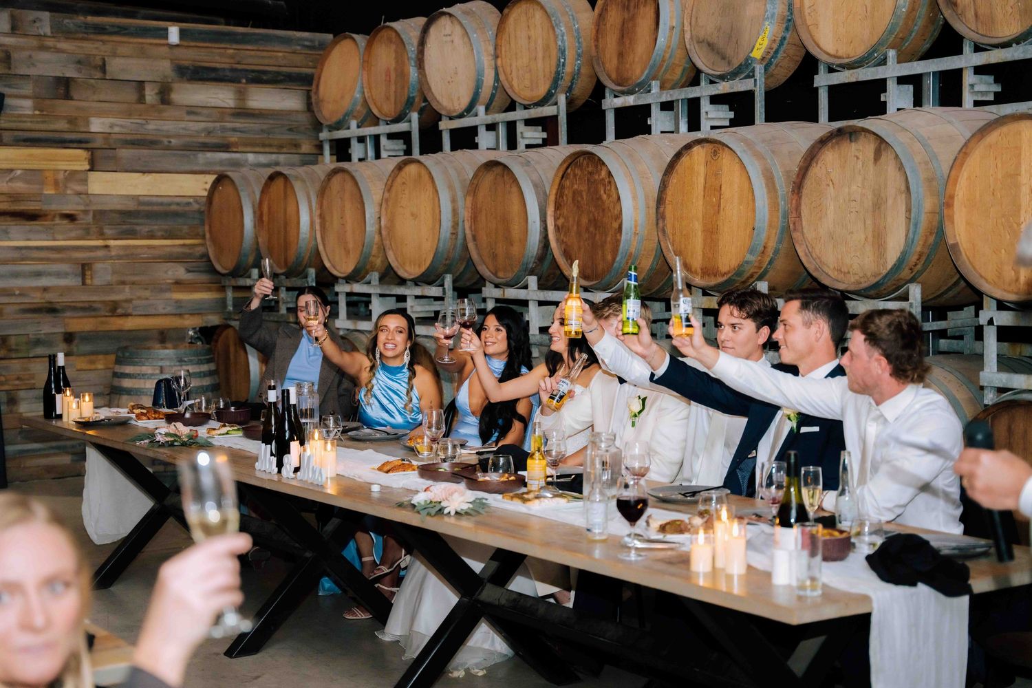 Wedding party raises champagne glasses in toast beside oak barrels at rustic winery reception dinner.