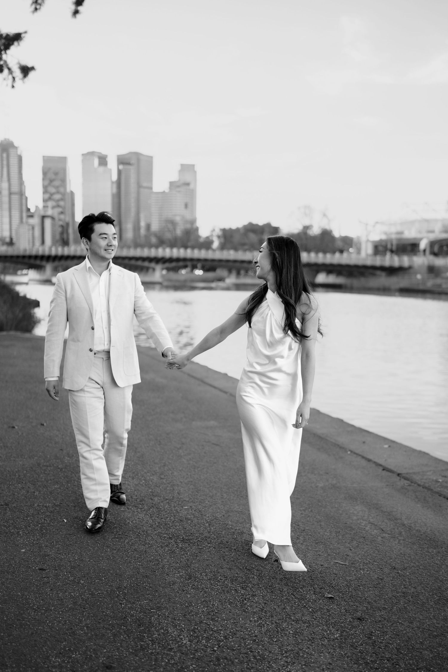 Black and white photo of Asian couple in white outfits holding hands while walking along a riverside path.