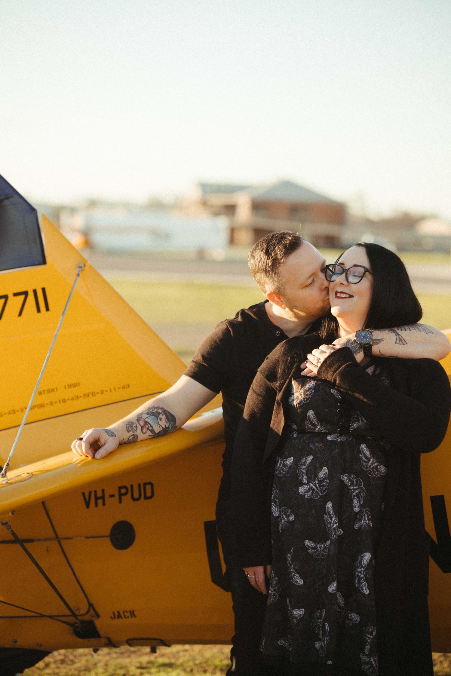 Man kissing woman on cheek beside a yellow vintage biplane VH-PUD at golden hour.