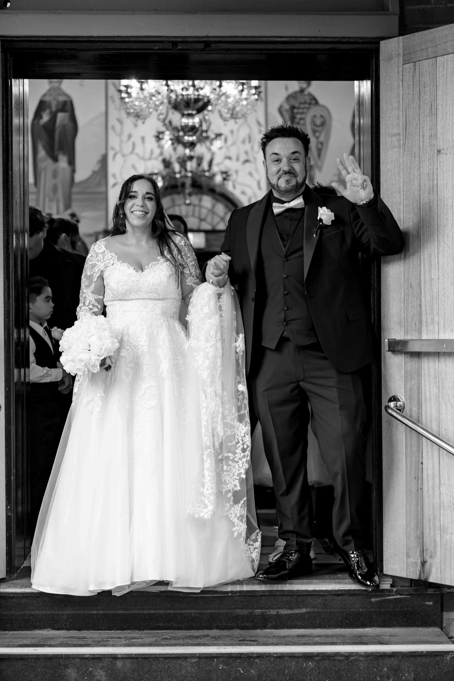 Joyful newlyweds exit church doors hand in hand after wedding ceremony, black and white.