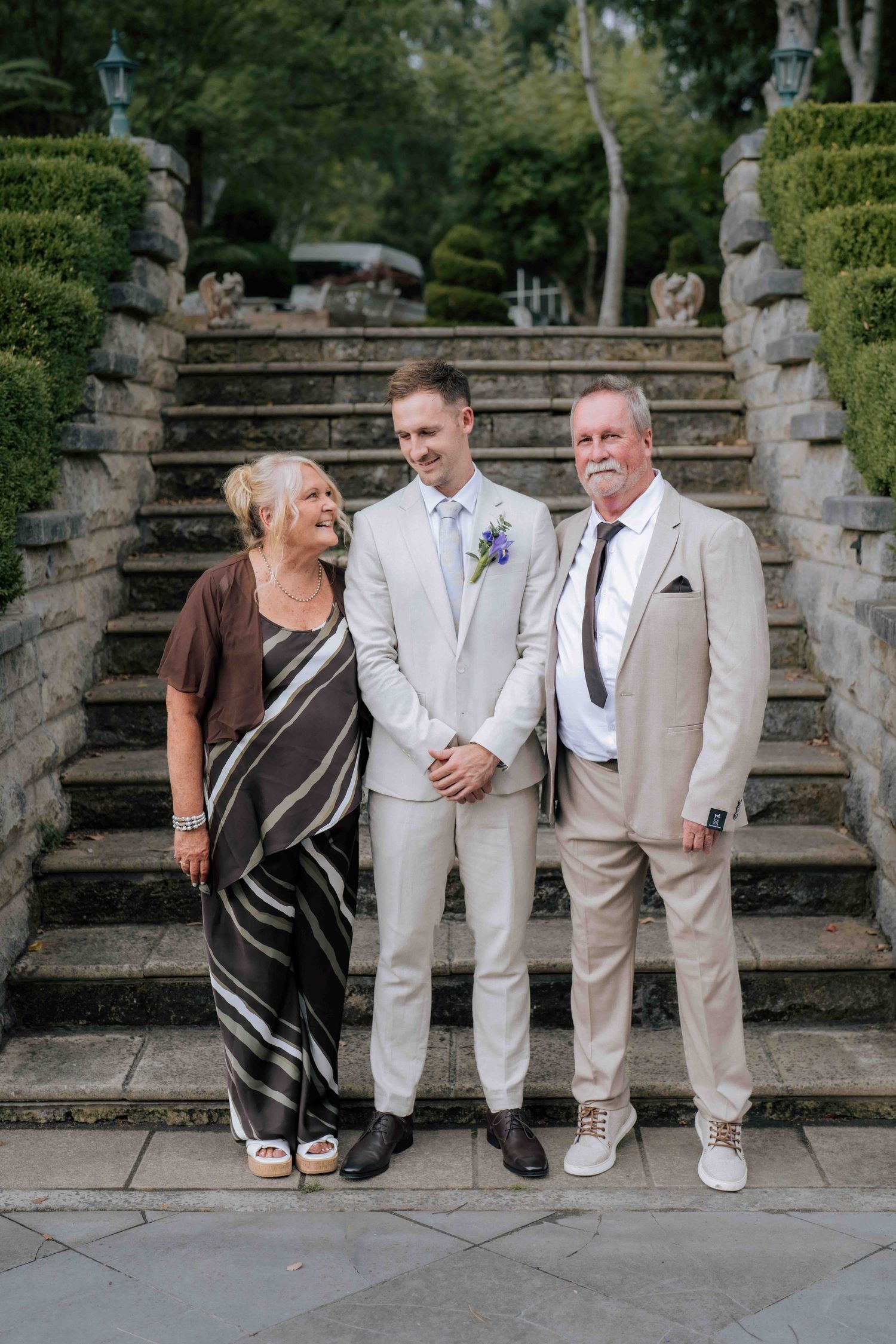 Groom in cream suit smiling with his parents on stone steps at an outdoor wedding venue.
