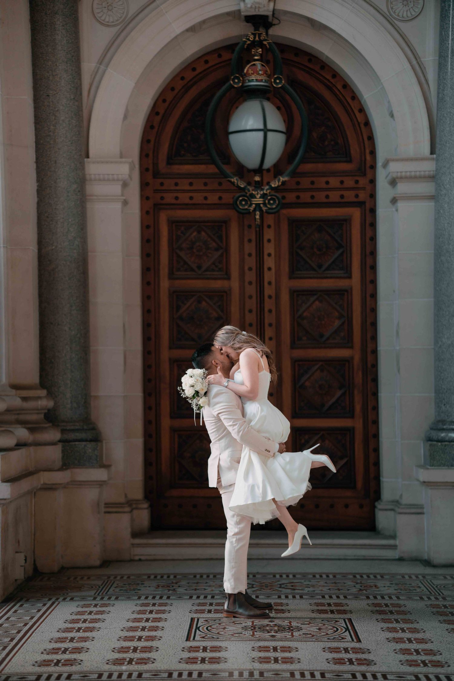 Groom lifting bride in a joyful embrace before an ornate wooden arched door.
