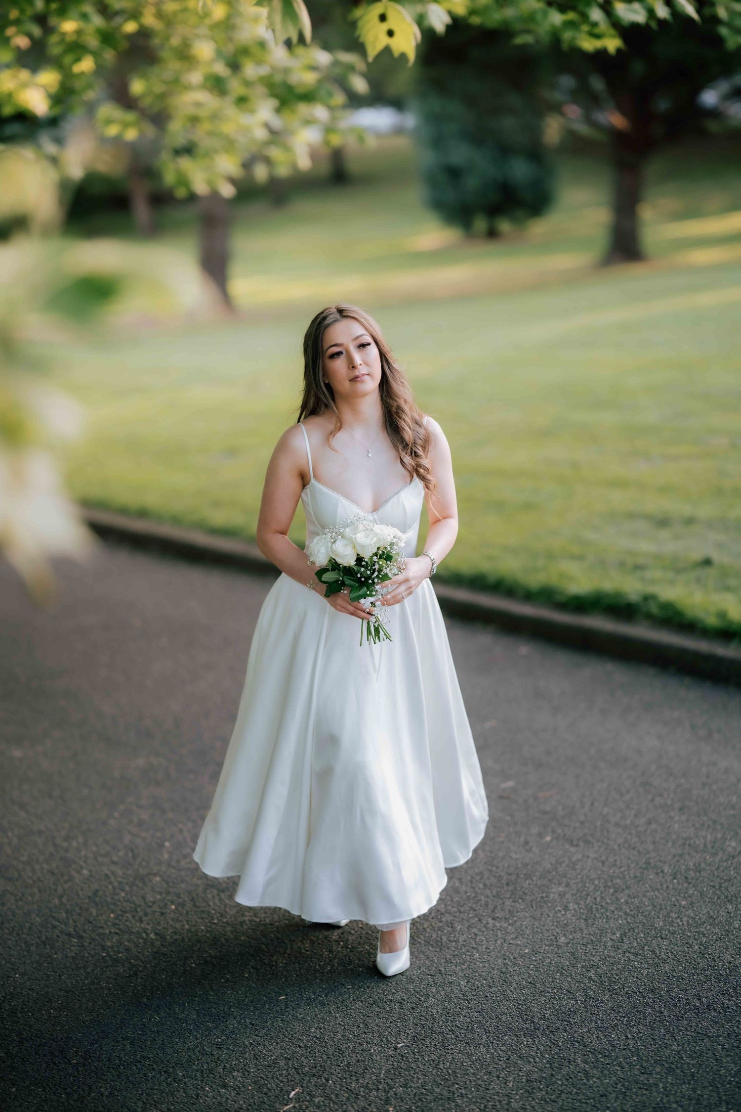 Bride in white tea-length gown holding white rose bouquet walking on park pathway at dusk.