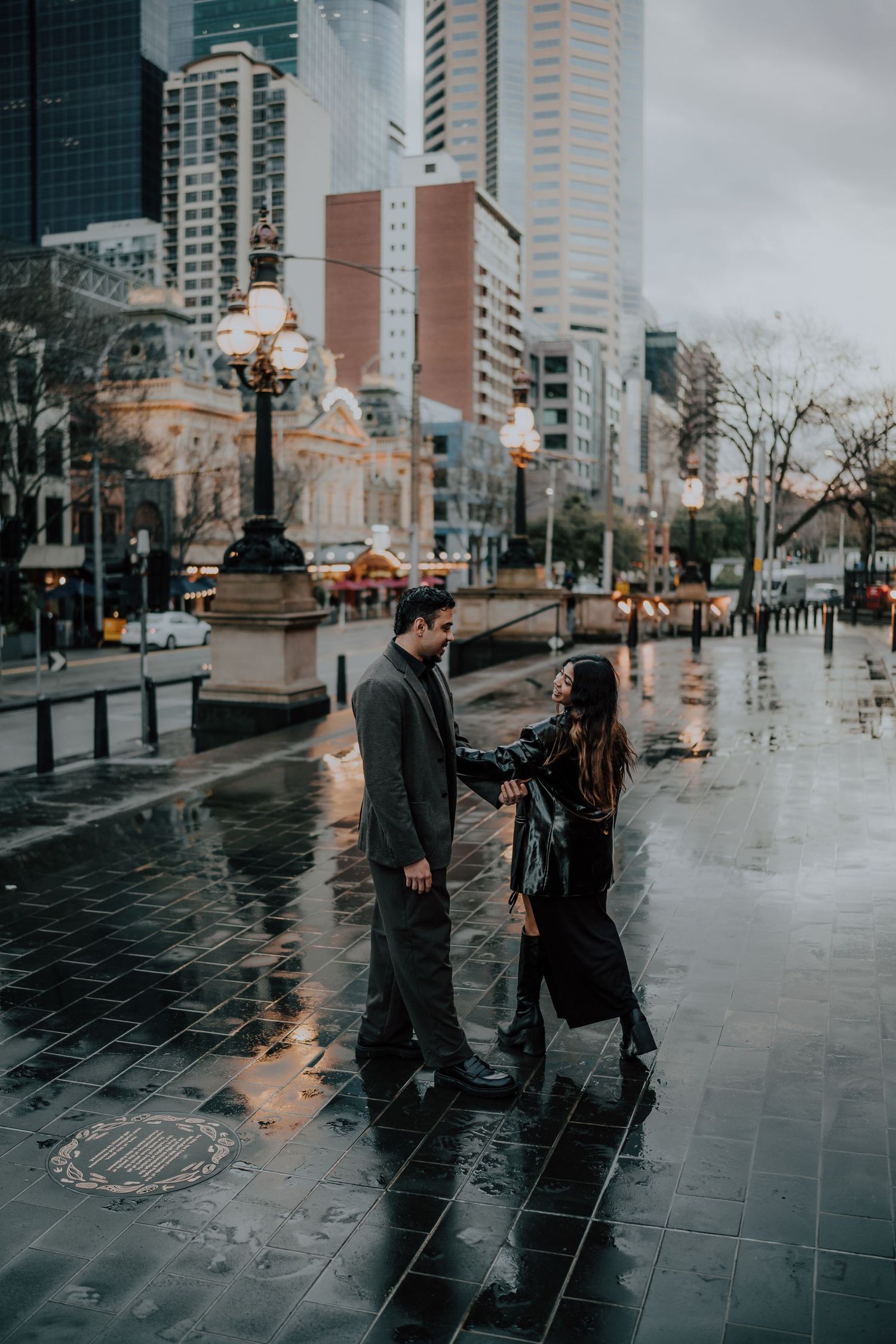 Couple holding hands walking on rain-soaked city streets with glowing lamp posts and skyscrapers in background.