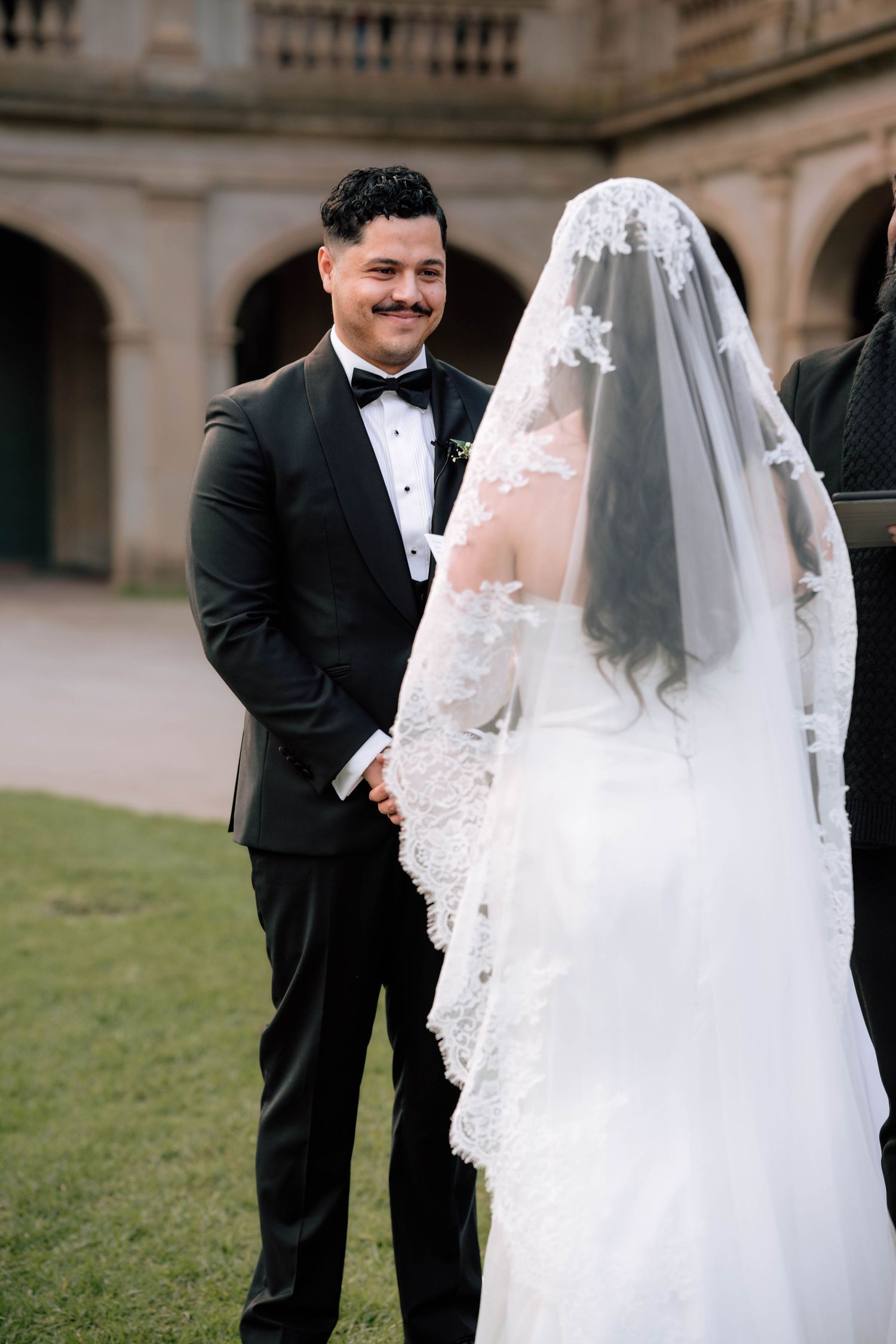Groom in black tuxedo smiles warmly at bride wearing ornate lace cathedral veil during ceremony.