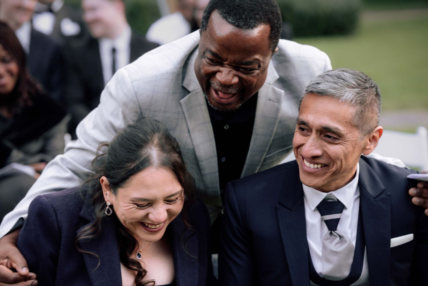 Three wedding guests laughing and embracing joyfully during outdoor wedding ceremony.