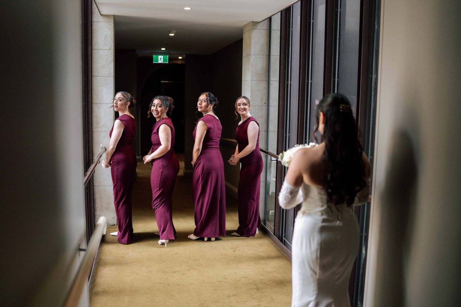 Bride in white gown viewing four bridesmaids in burgundy dresses lined up in a hotel hallway.